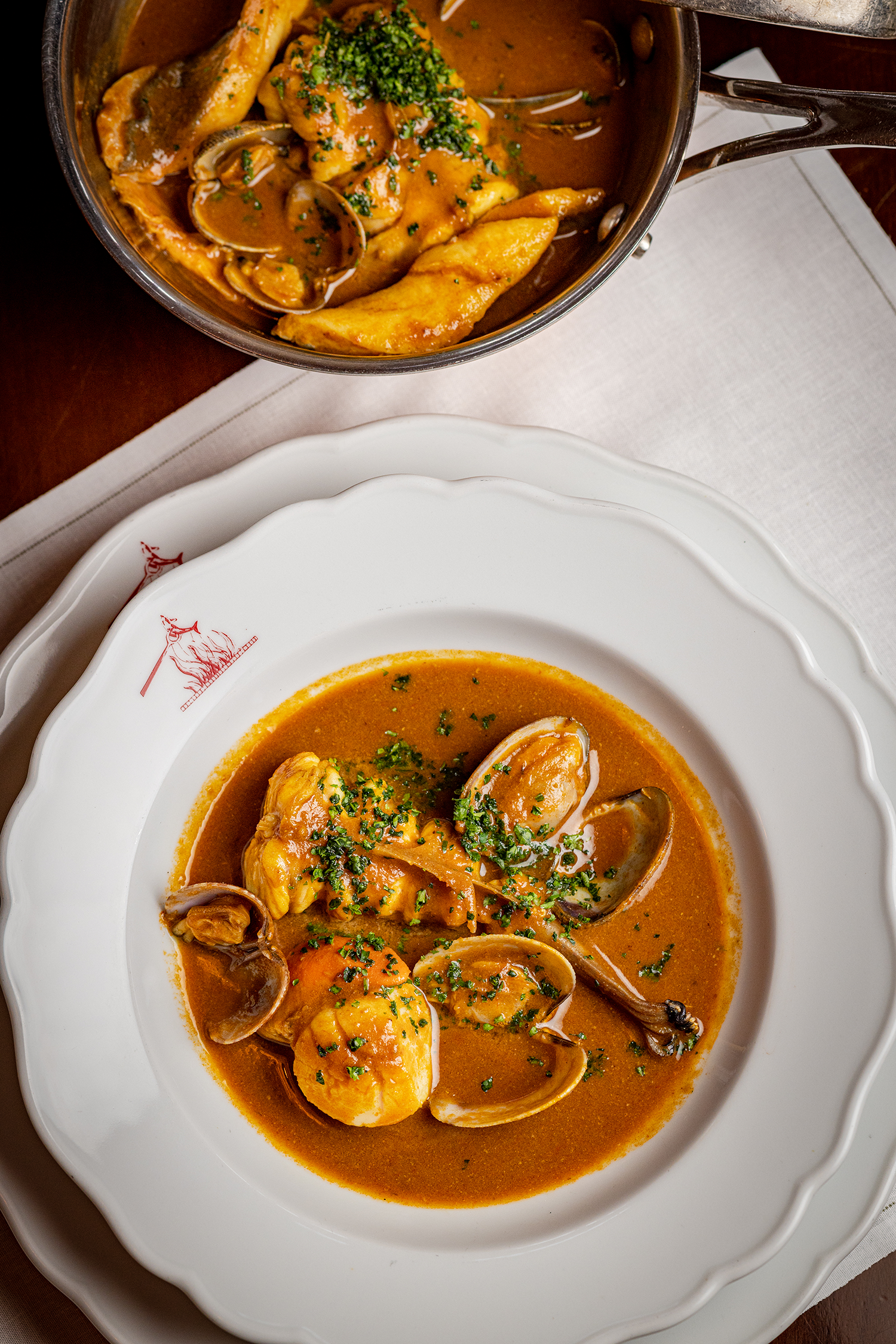 A bowl of seafood stew with clams, chicken, vegetables, and garnished with chopped herbs on a white plate with a decorative edge.