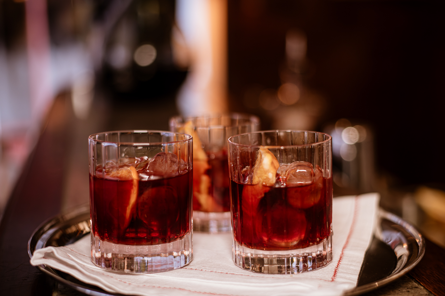 Three glasses of red cocktails with ice and orange slices on a silver tray with a white napkin.