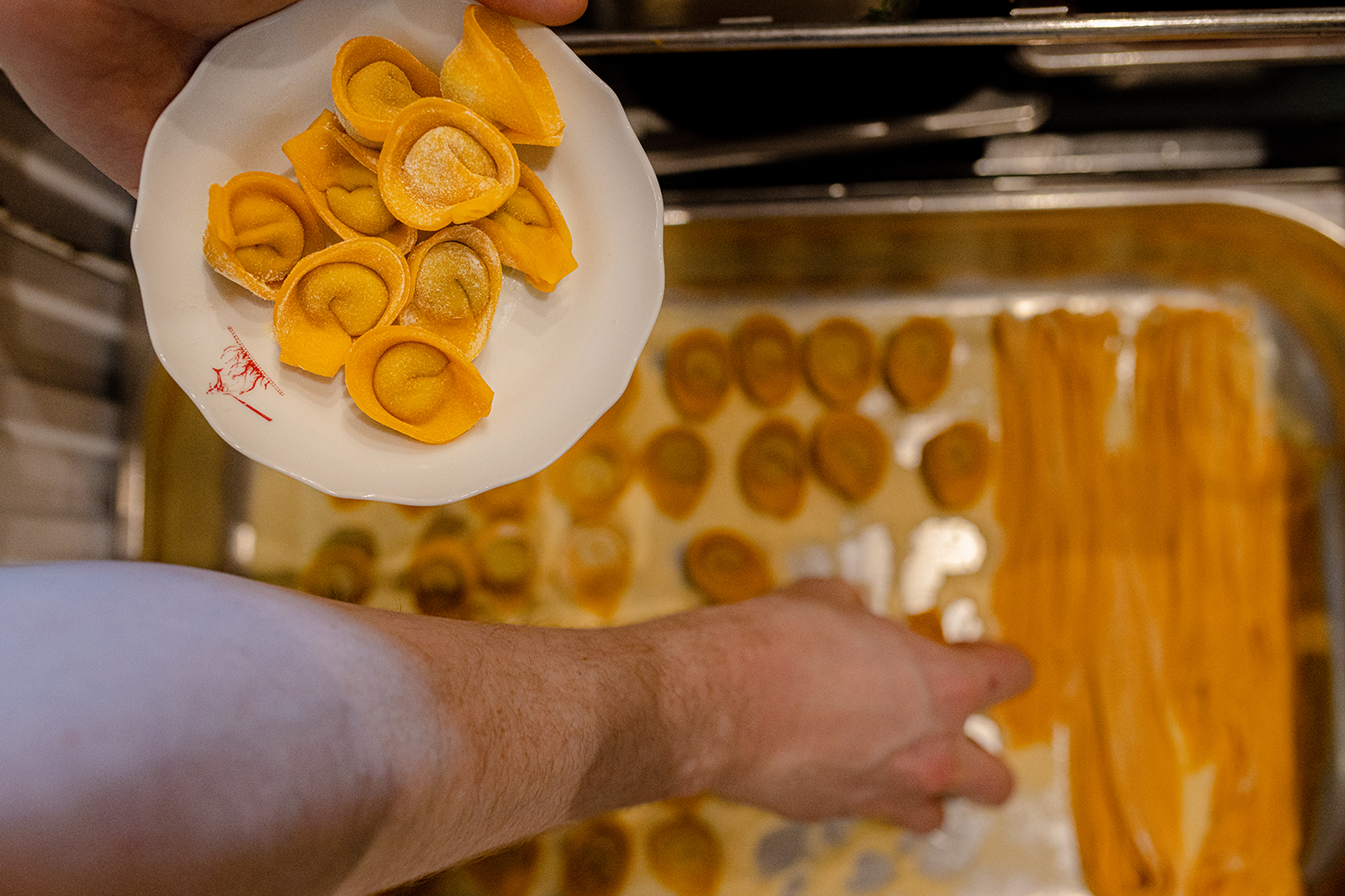 A person is holding a small white plate with yellow tortellini pasta. In the background, there is a baking tray with more tortellini and rolled-out dough.