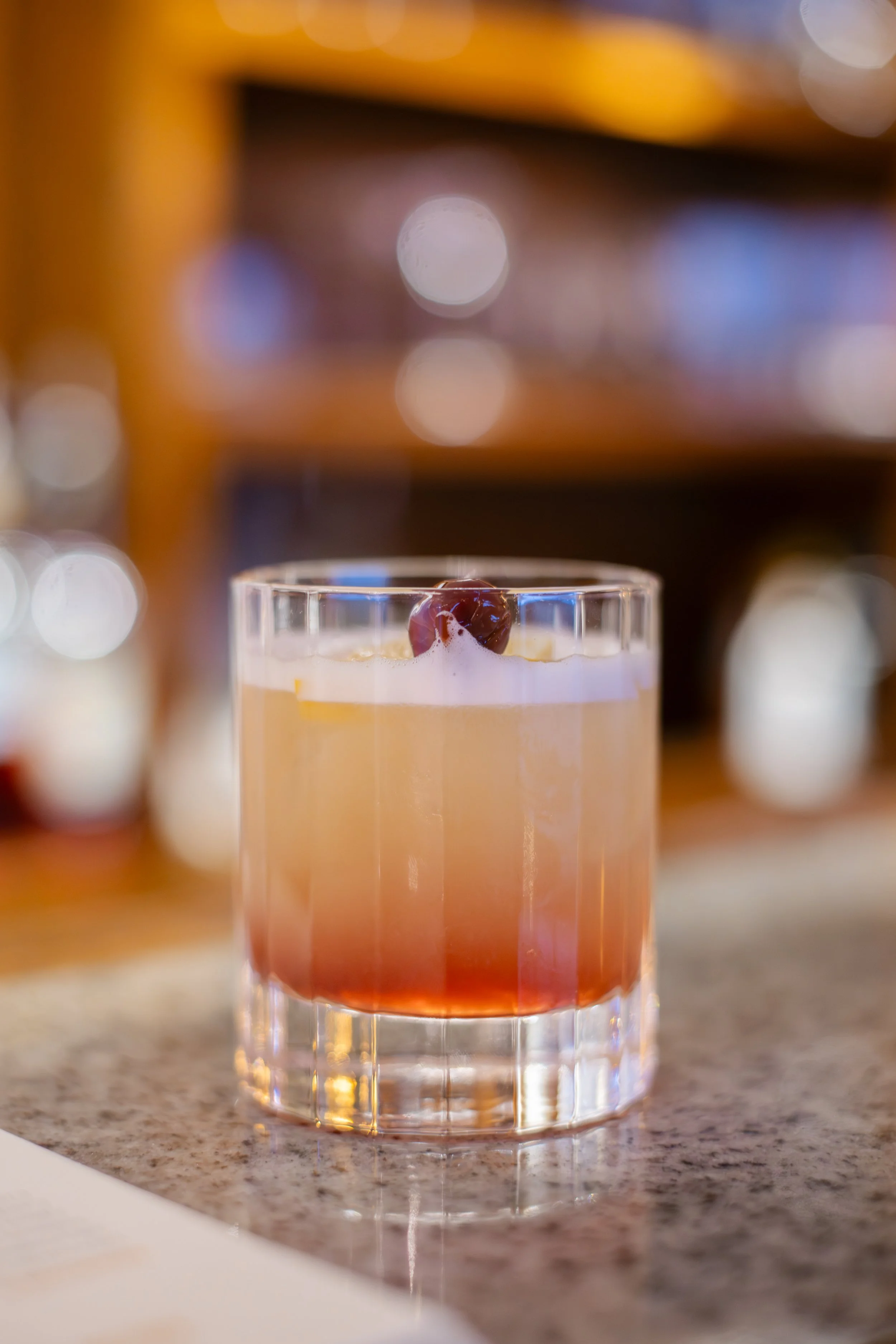 A glass of layered cocktail with a cherry on top, placed on a granite countertop with a blurred background featuring bokeh lights.