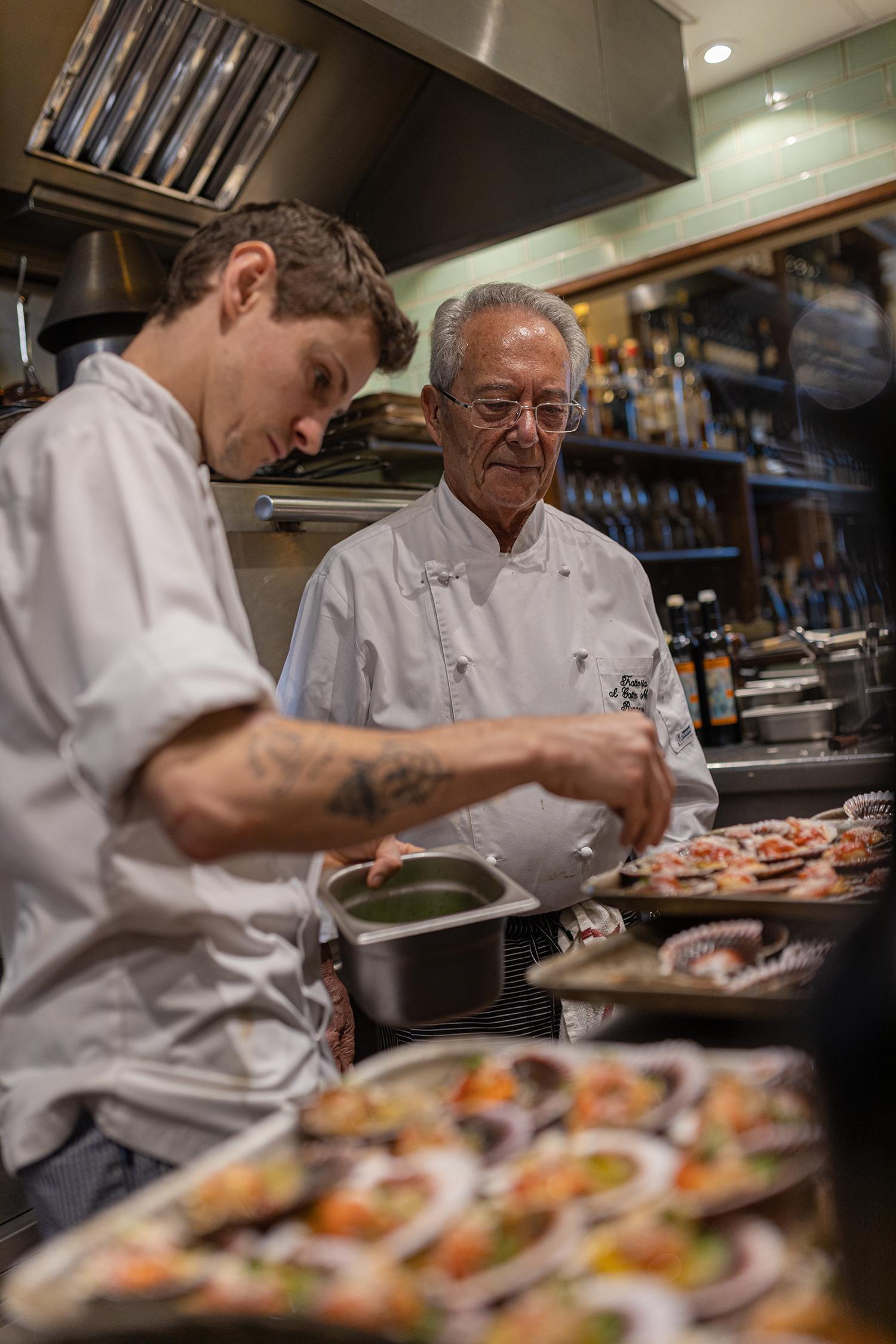 Two chefs in a kitchen preparing food, with a plate of assorted sushi in the foreground.