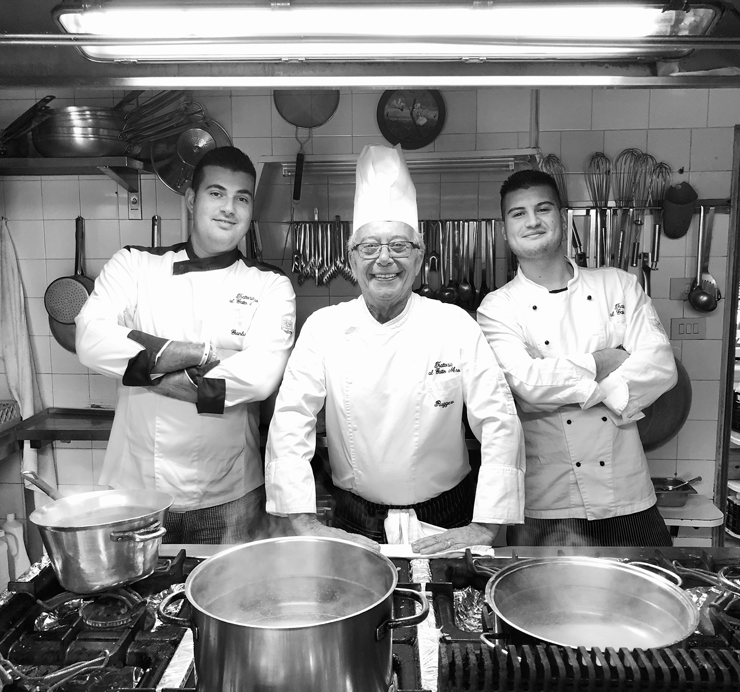 Three chefs in a kitchen, two young men and an older man, all wearing chef uniforms, standing in front of cooking pots and kitchen utensils.