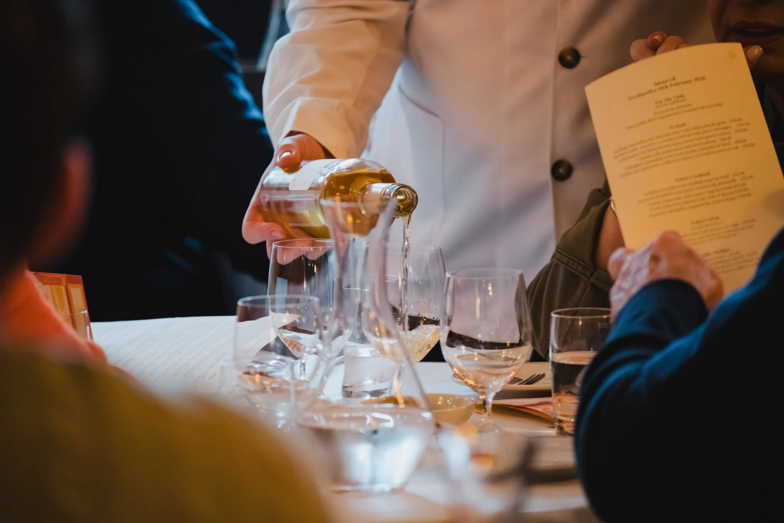 A chef pouring honey or syrup into glasses at a dining table during a tasting or food event.