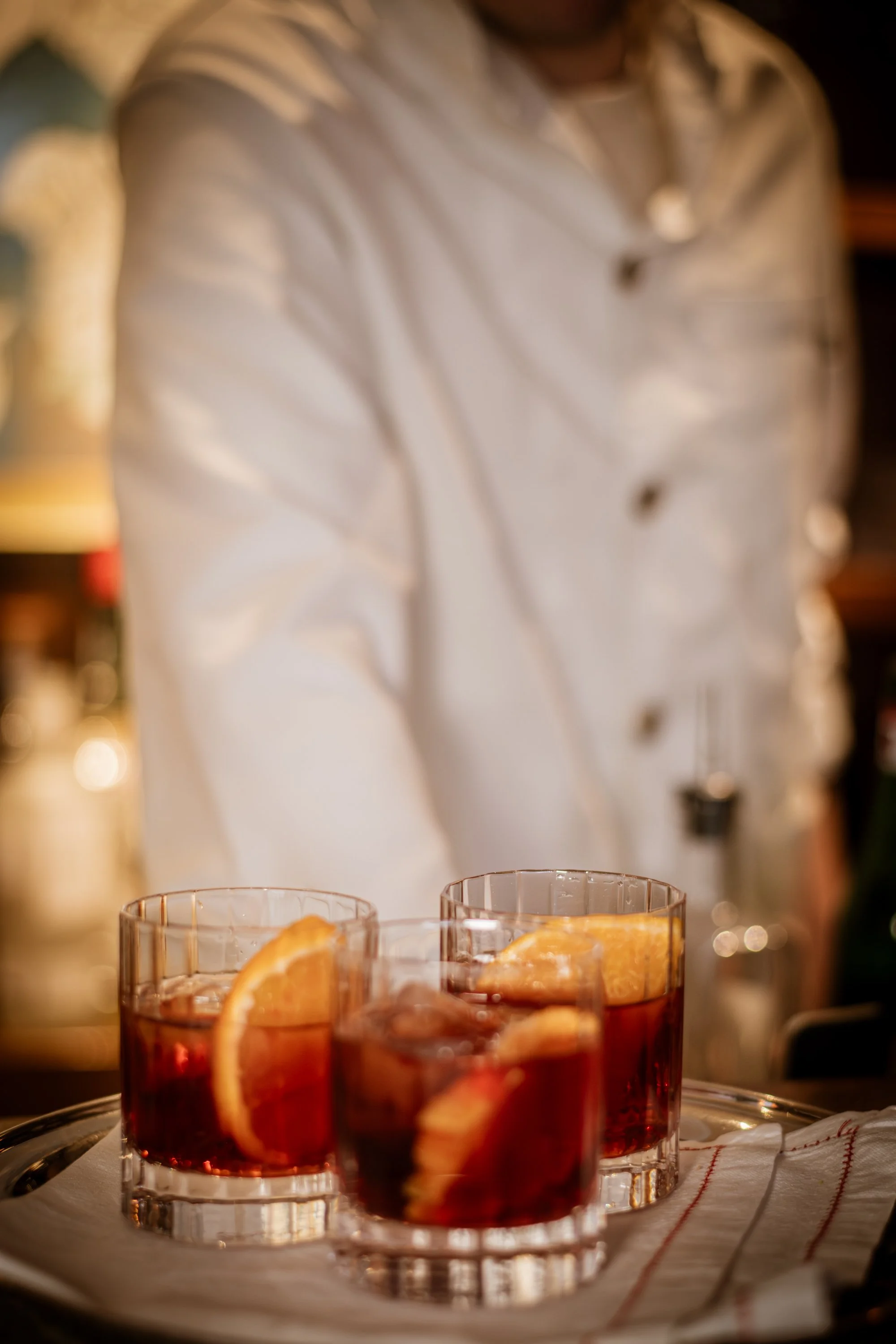 Three glasses of a red beverage with orange slices inside, placed on a tray on a table with a white cloth. A person dressed as a chef or bartender is blurred in the background.