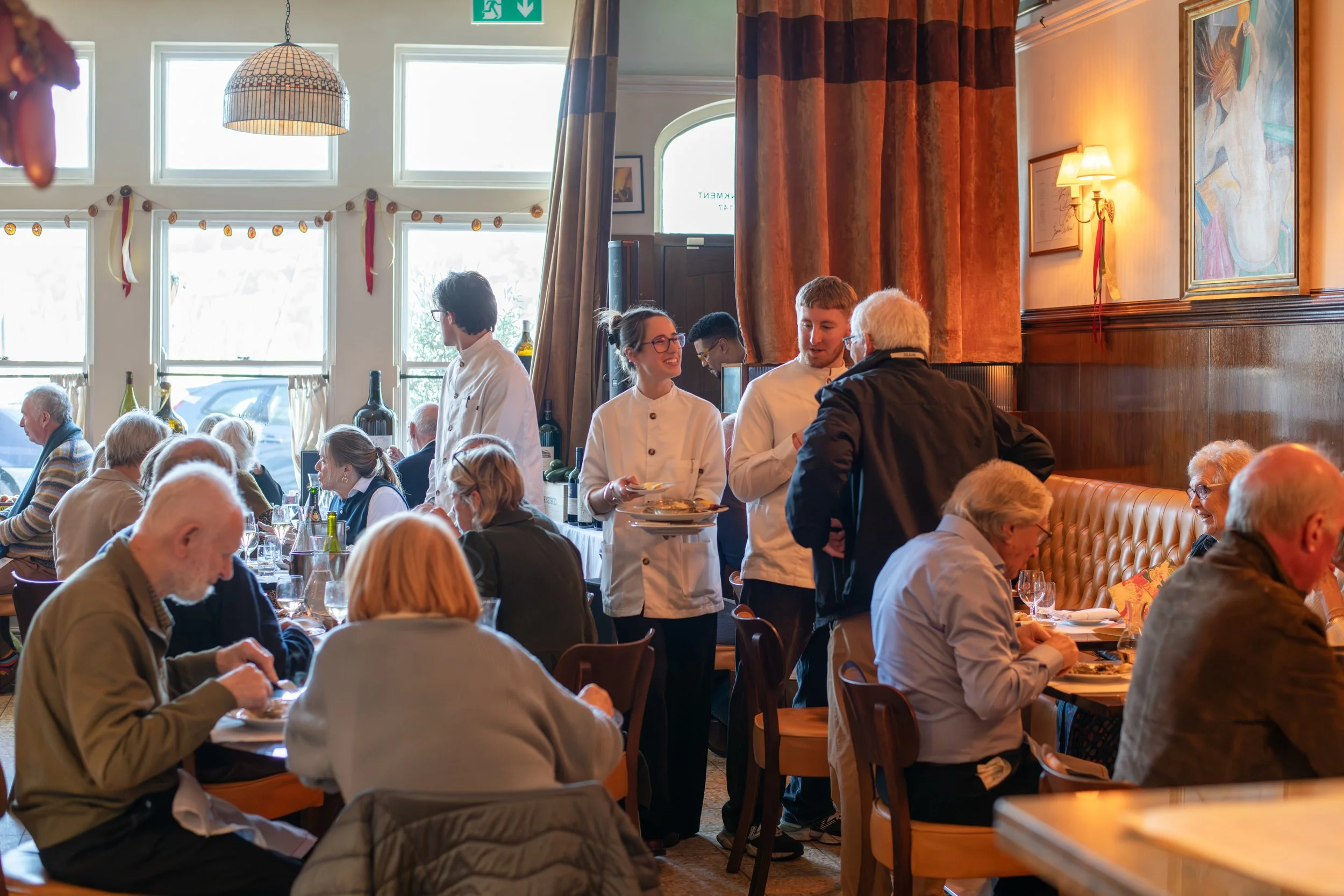 A busy restaurant with several diners, waitstaff serving food, and a warm, inviting interior with large windows, curtains, and wall art.