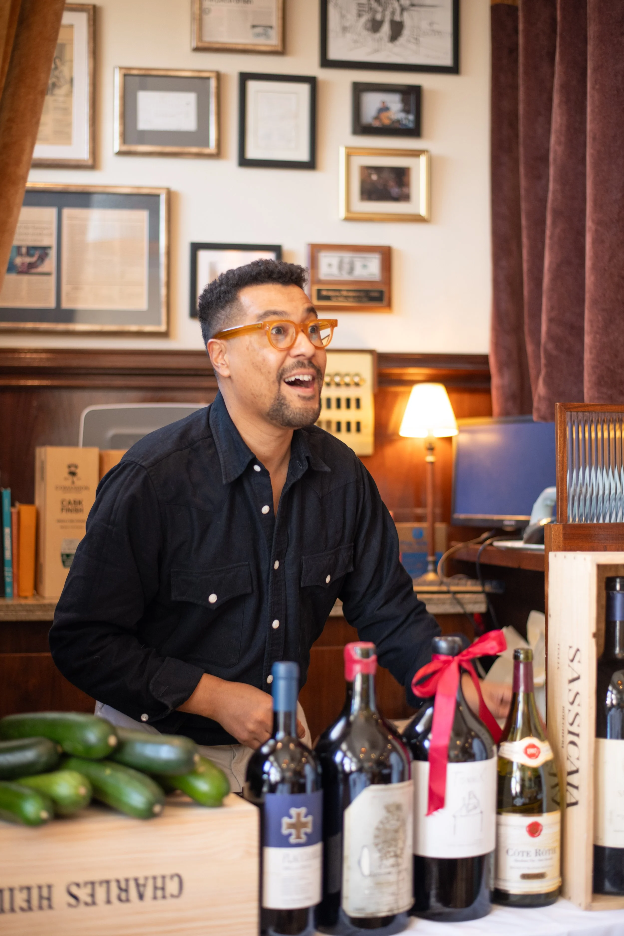 A man with glasses and a black shirt smiling at a market or winery table with bottles of wine, fresh vegetables, and wine boxes. The background features a wall decorated with framed pictures and certificates, warm lighting, and red curtains.