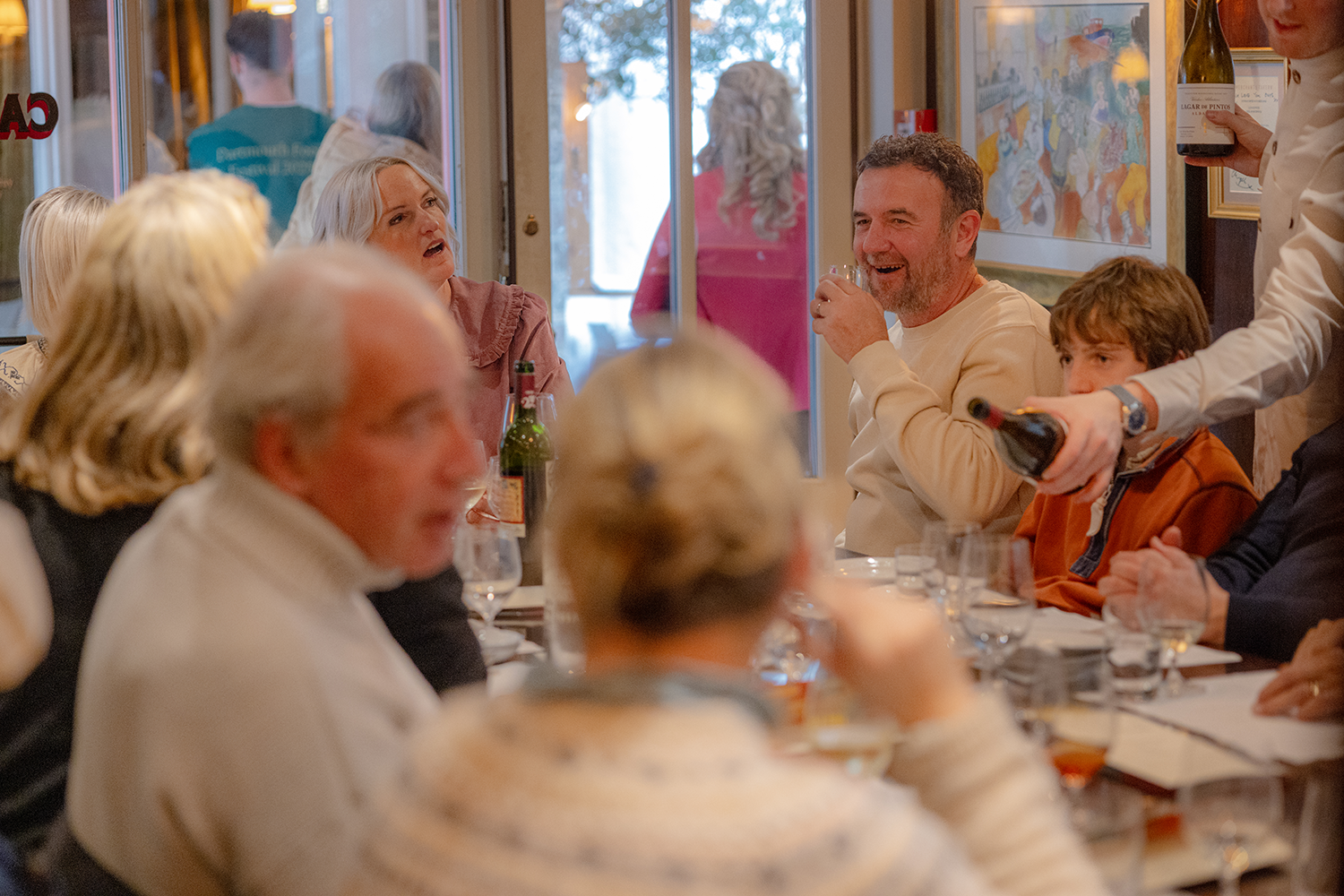 People gathered at a dinner table in a restaurant, enjoying drinks and conversation, with some standing and serving beverages.