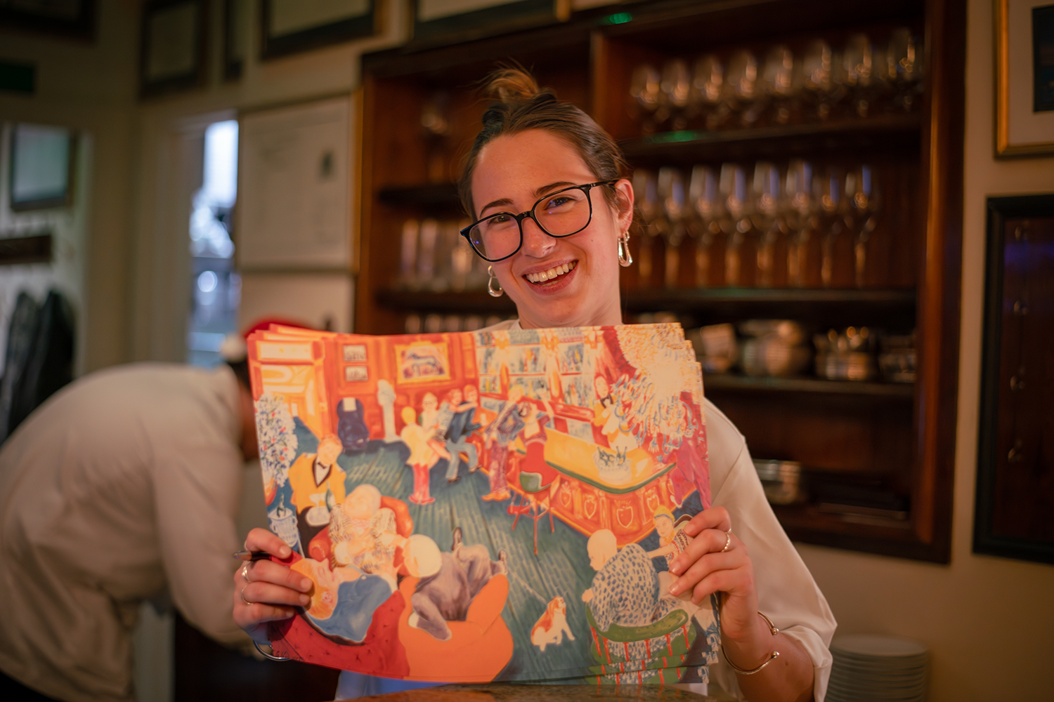 A woman with glasses and earrings smiling and holding a colorful painting of a lively indoor gathering with people, a dog, and artwork on the walls.