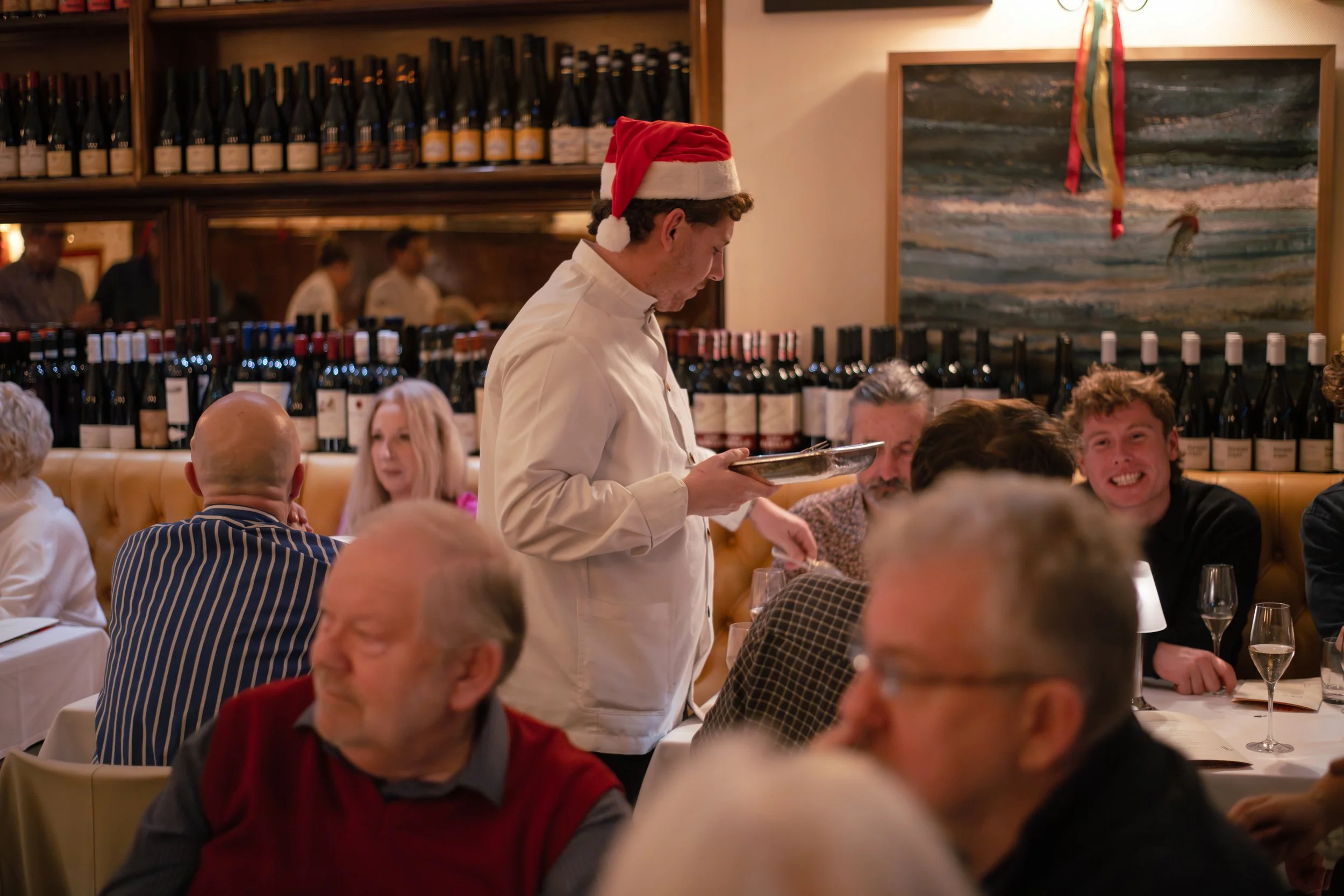 A restaurant scene with multiple diners seated at tables. A waiter wearing a Santa hat and white shirt is serving food. The background shows a wine display on shelves and a painting on the wall with holiday decorations.