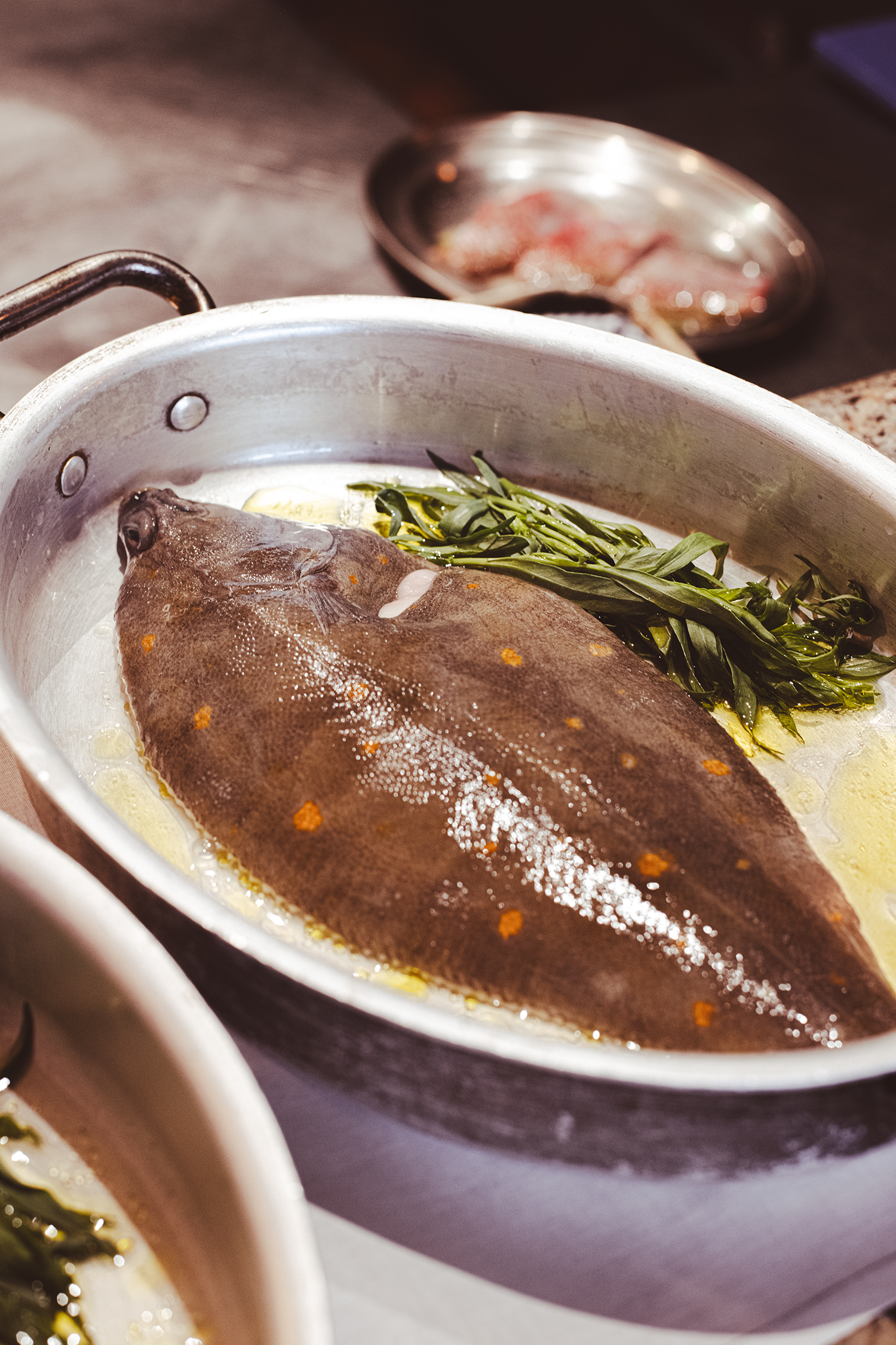 A fish, possibly a trout or similar species, rests in a metal pan with herb sprigs. In the background, there is a small metal dish with raw meat pieces.