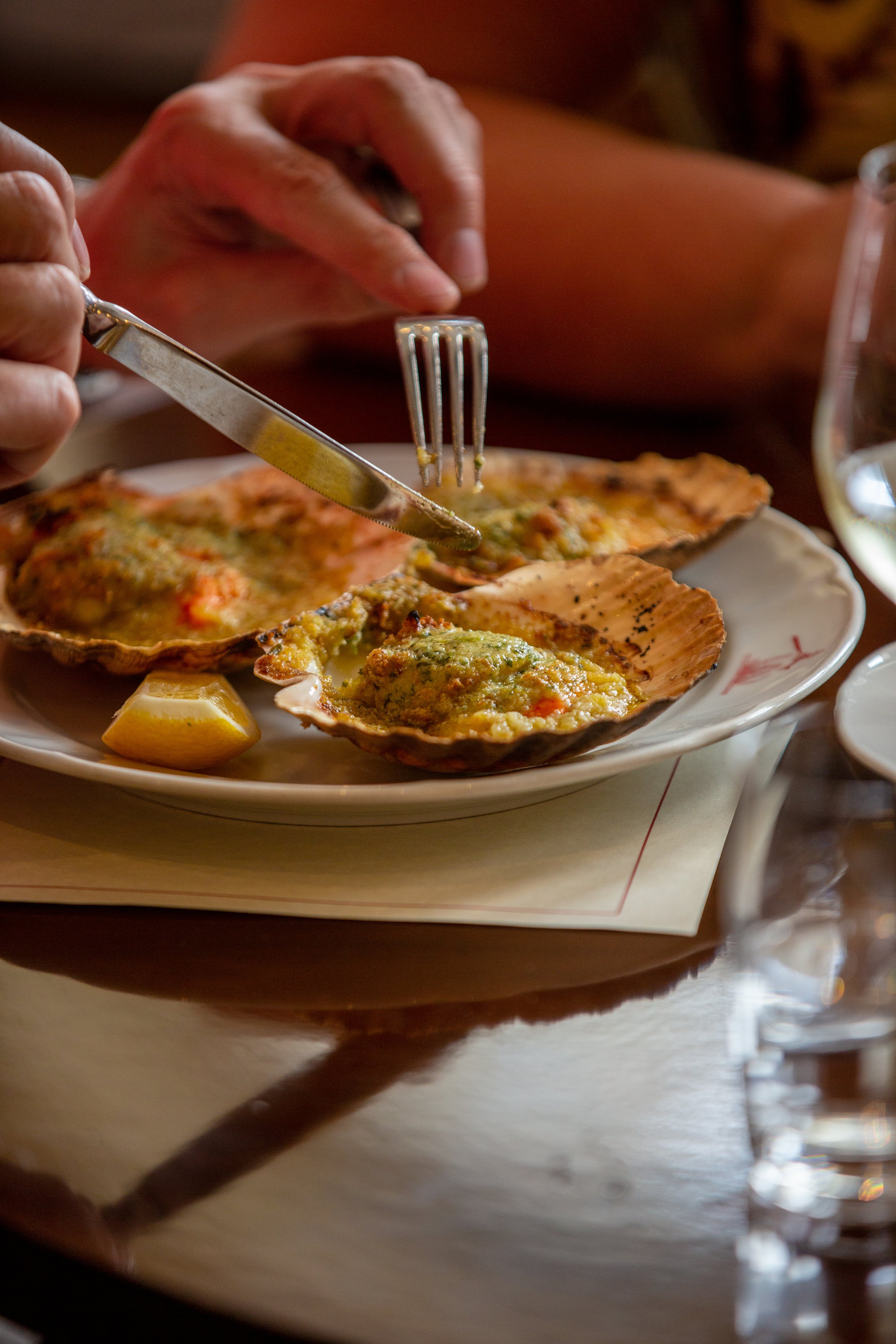 A close-up of a person's hand using a fork and knife to cut into a baked oyster on a plate, with lemon wedges and a glass of water nearby.