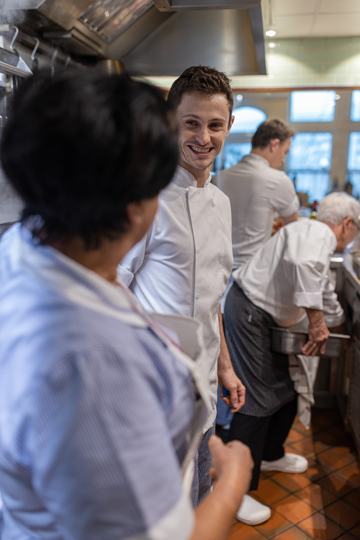Group of chefs in a kitchen, smiling and preparing food, wearing white chef uniforms.