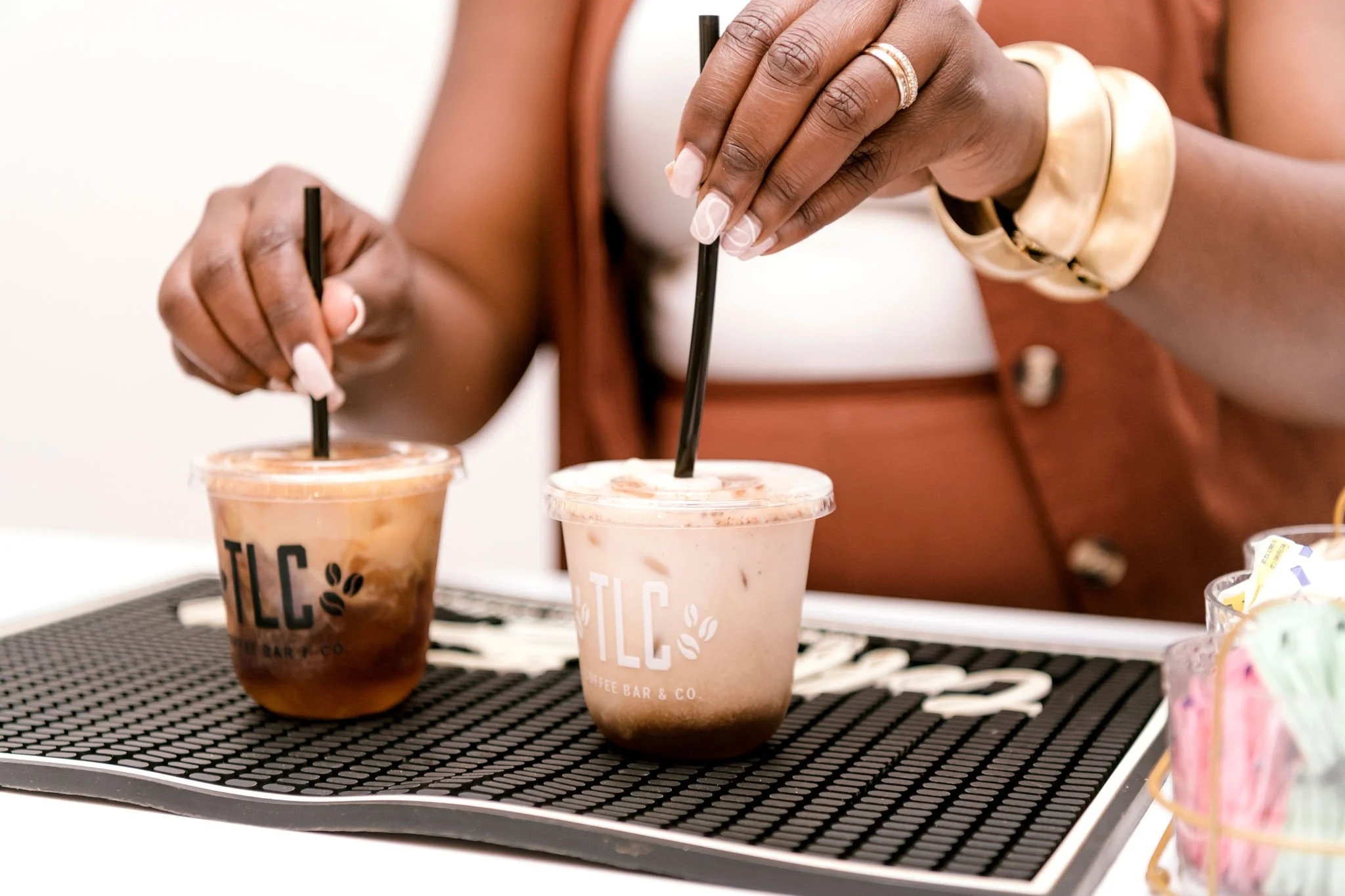 Two people holding iced coffee drinks with black straws at a coffee shop, with TLC logo on the cups.