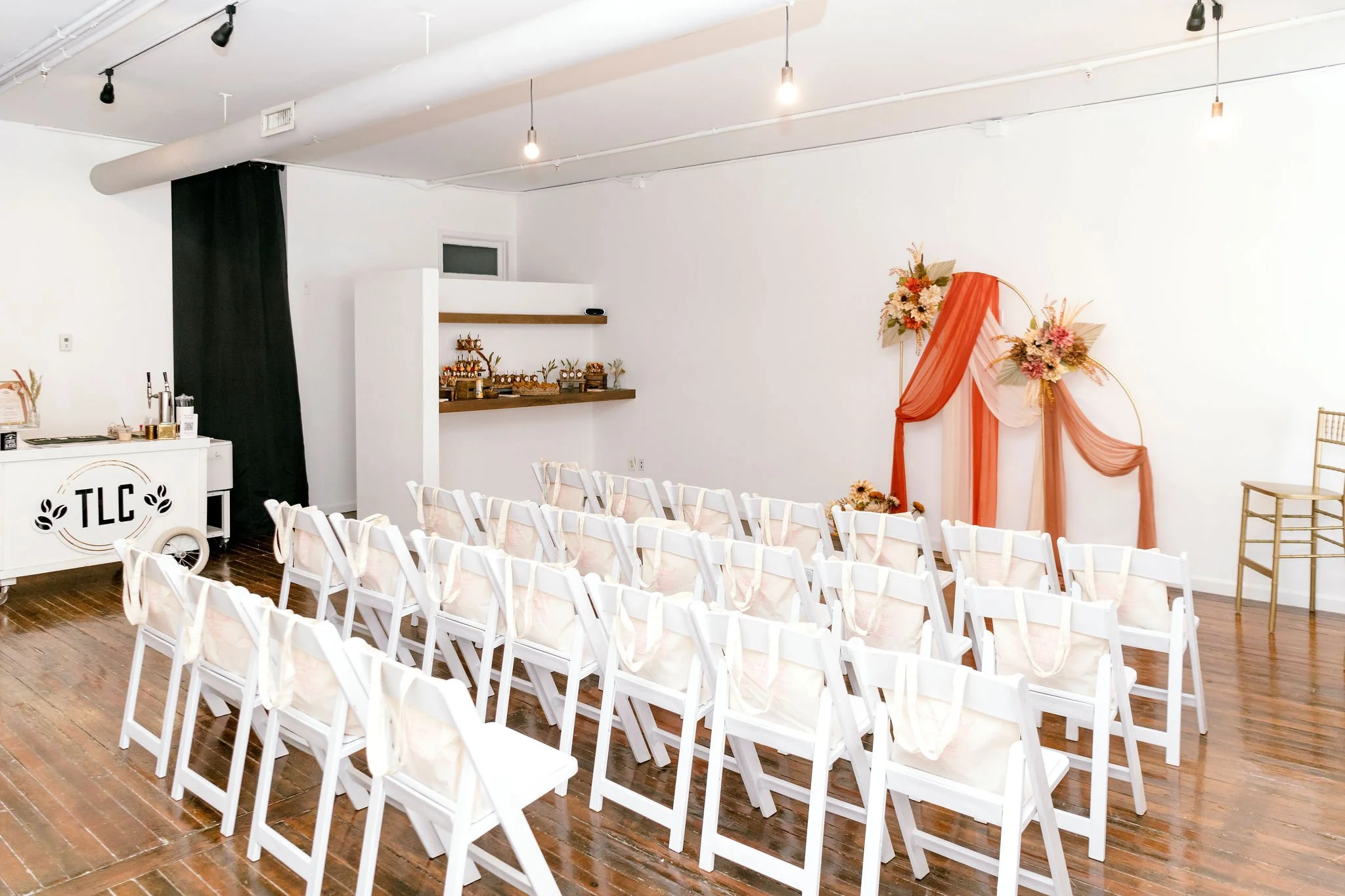 Indoor wedding ceremony setup with white chairs, floral decorations, and a backdrop with orange and pink drapes.