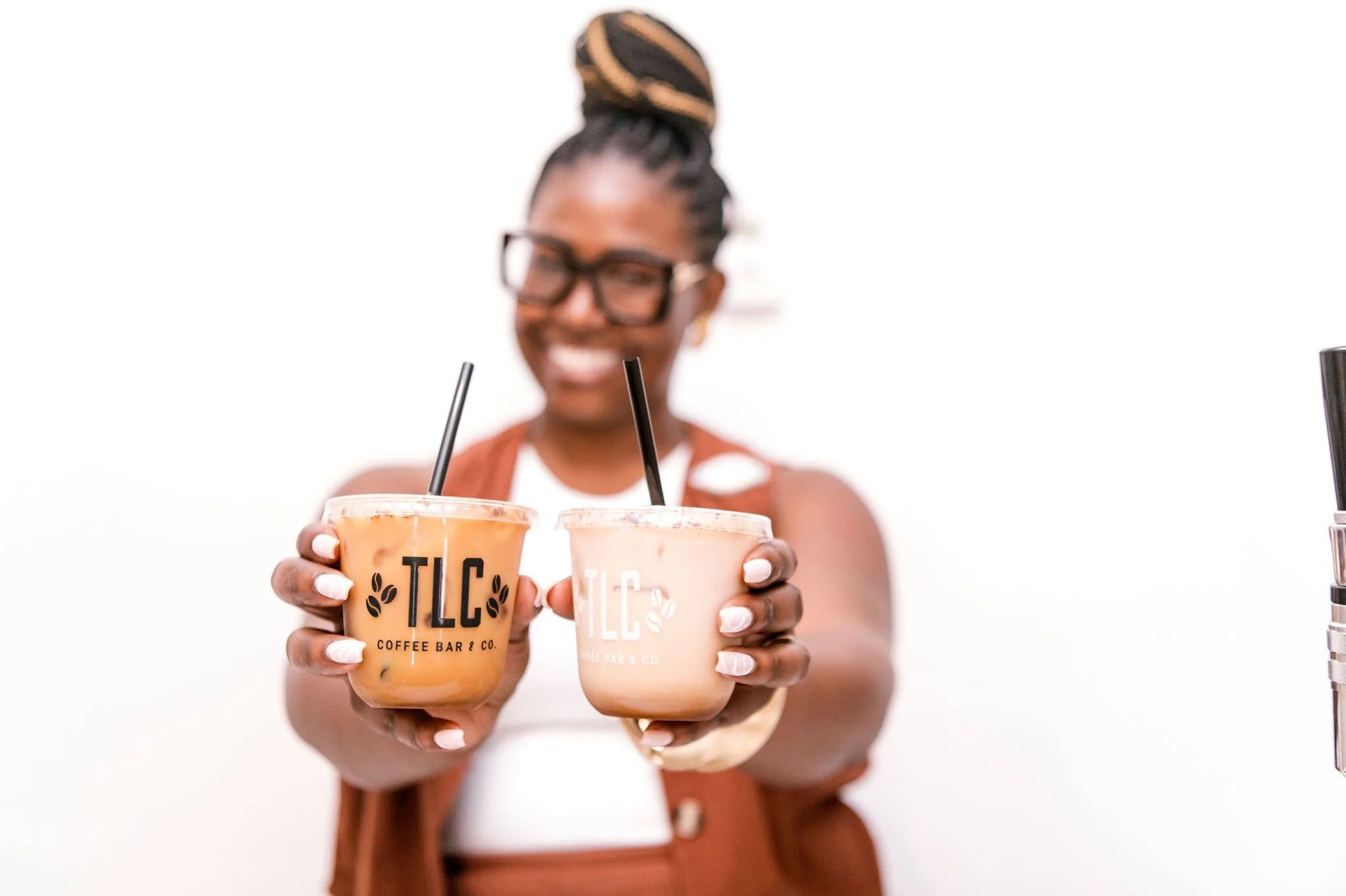A smiling woman with glasses holding two iced coffee drinks from TLC Coffee Bar & Co, with a white background