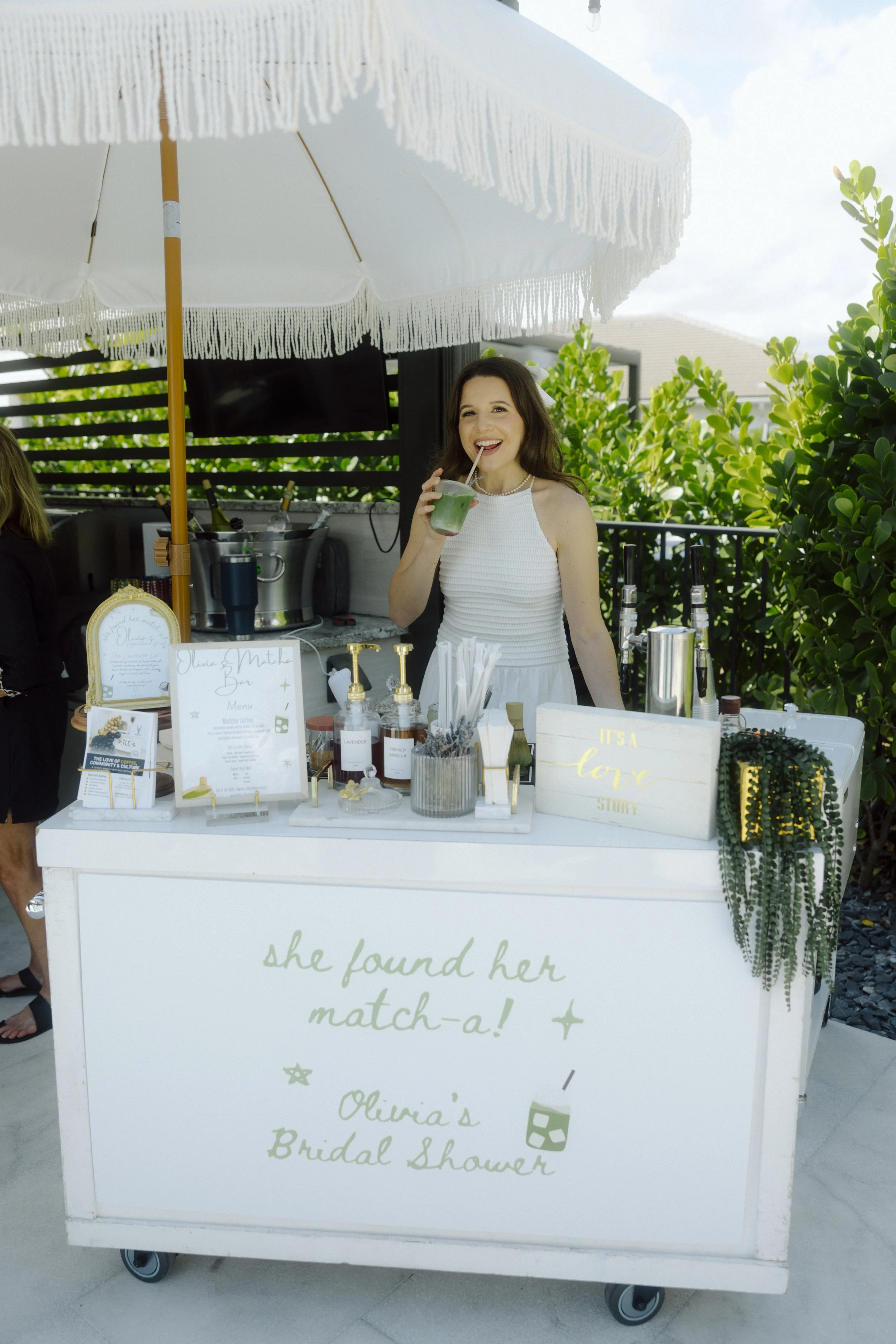 A woman smiling and drinking a green beverage through a straw at a bridal shower stand under a white umbrella. TLC Coffee Bar & Co. Premium Mobile Café Bar Experience for every event serving all of South Florida. Specialty Coffee, Matcha, and Teas.