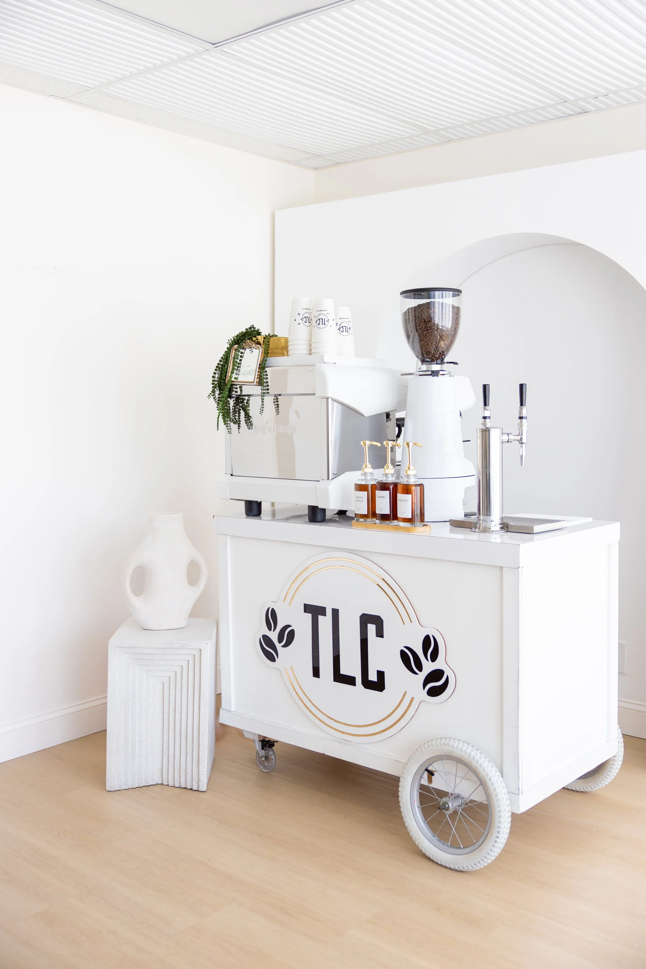 A white coffee station with the logo 'TLC' on the front, featuring a coffee grinder, brewing equipment, and soap dispensers, located against white walls with a light-colored wooden floor.