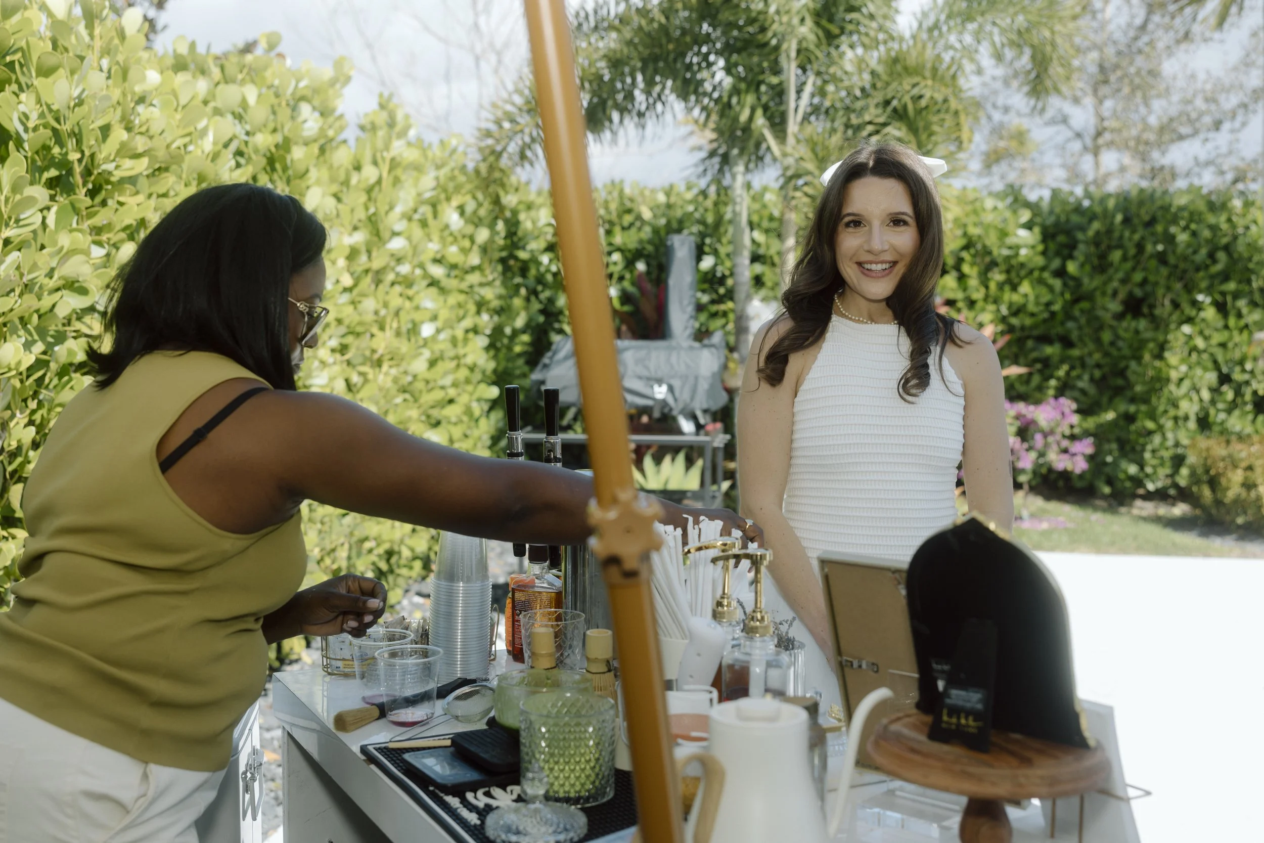 A smiling woman with long dark hair, wearing a white sleeveless top and a white bow in her hair, stands next to an outdoor beverage stand. A woman with dark skin and sunglasses, dressed in a yellow tank top and white pants, is preparing drinks. The s
