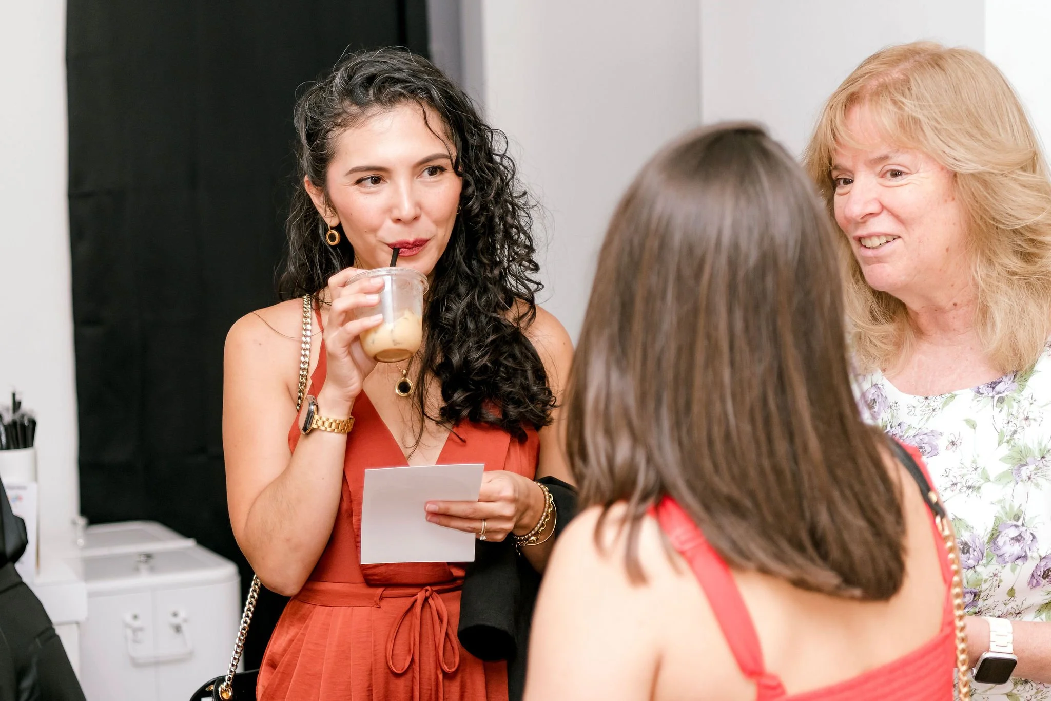 Three women engaged in conversation at a social event; one woman is holding a drink and looking at a card, another is smiling, and the third has her back to the camera.