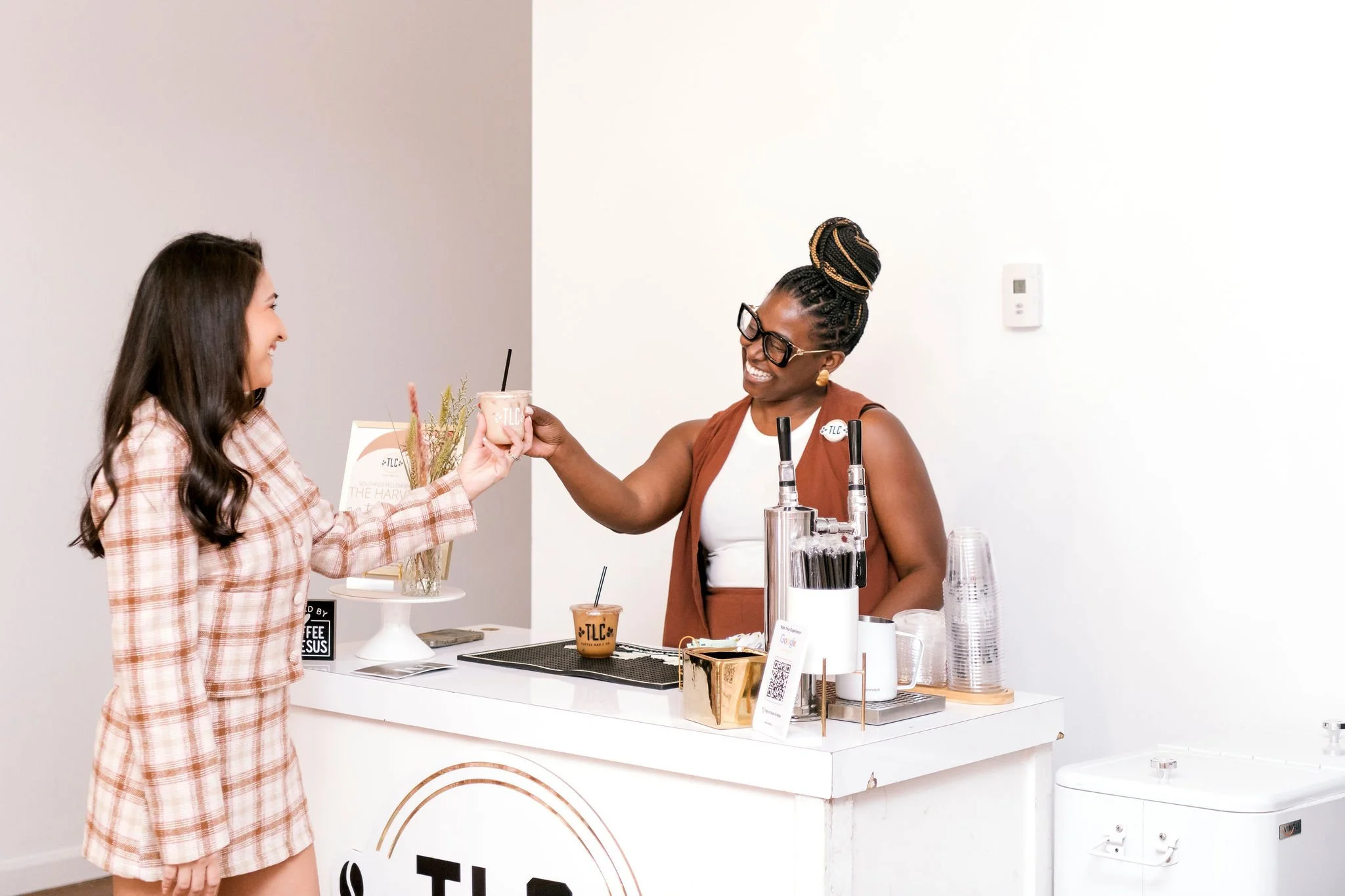 A woman with glasses and braided hair hands a coffee cup to a smiling customer at a coffee shop counter.