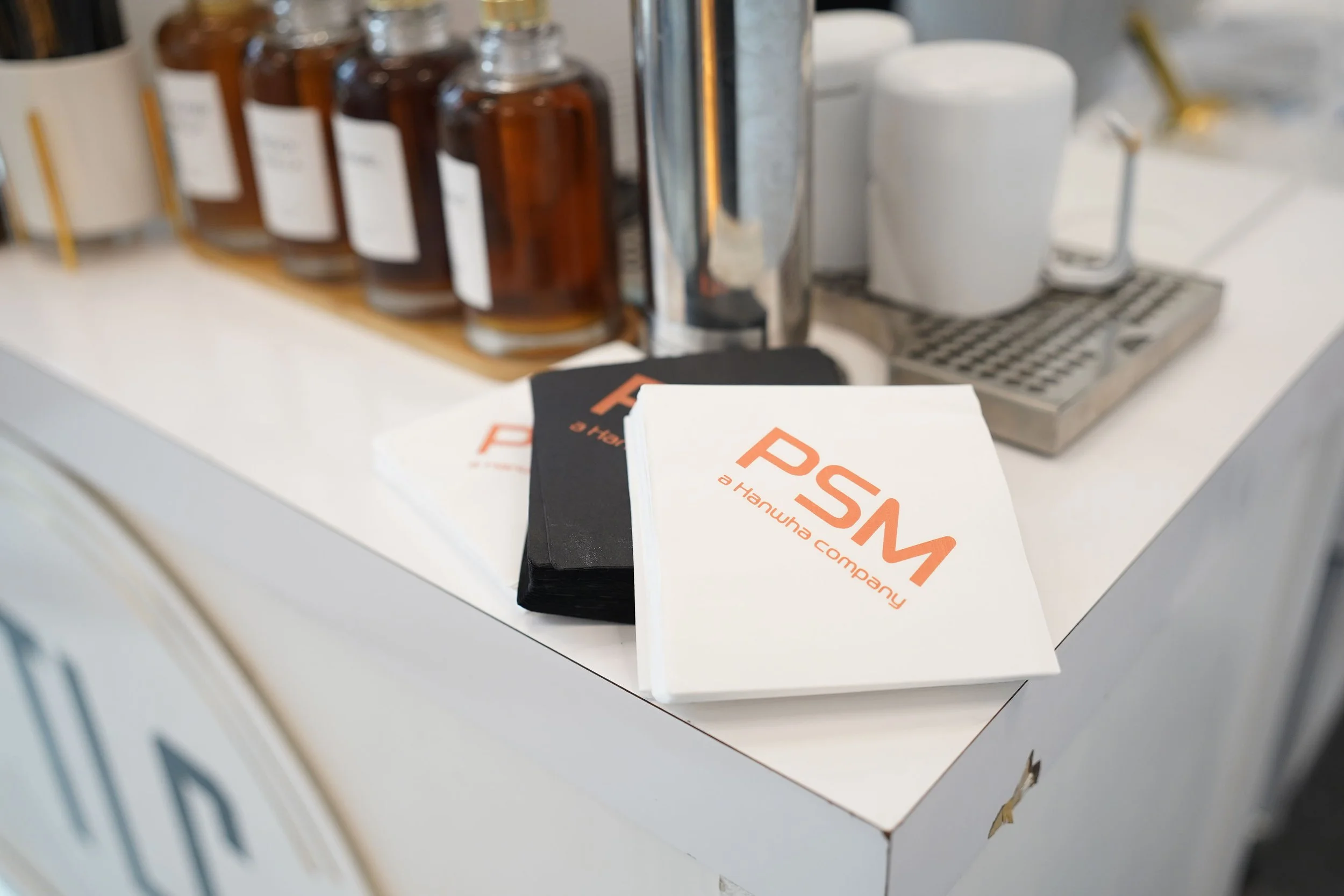 Stack of PTSM branded napkins on a white countertop in front of a coffee station with brown bottles, a stainless steel espresso machine, and a white cup.