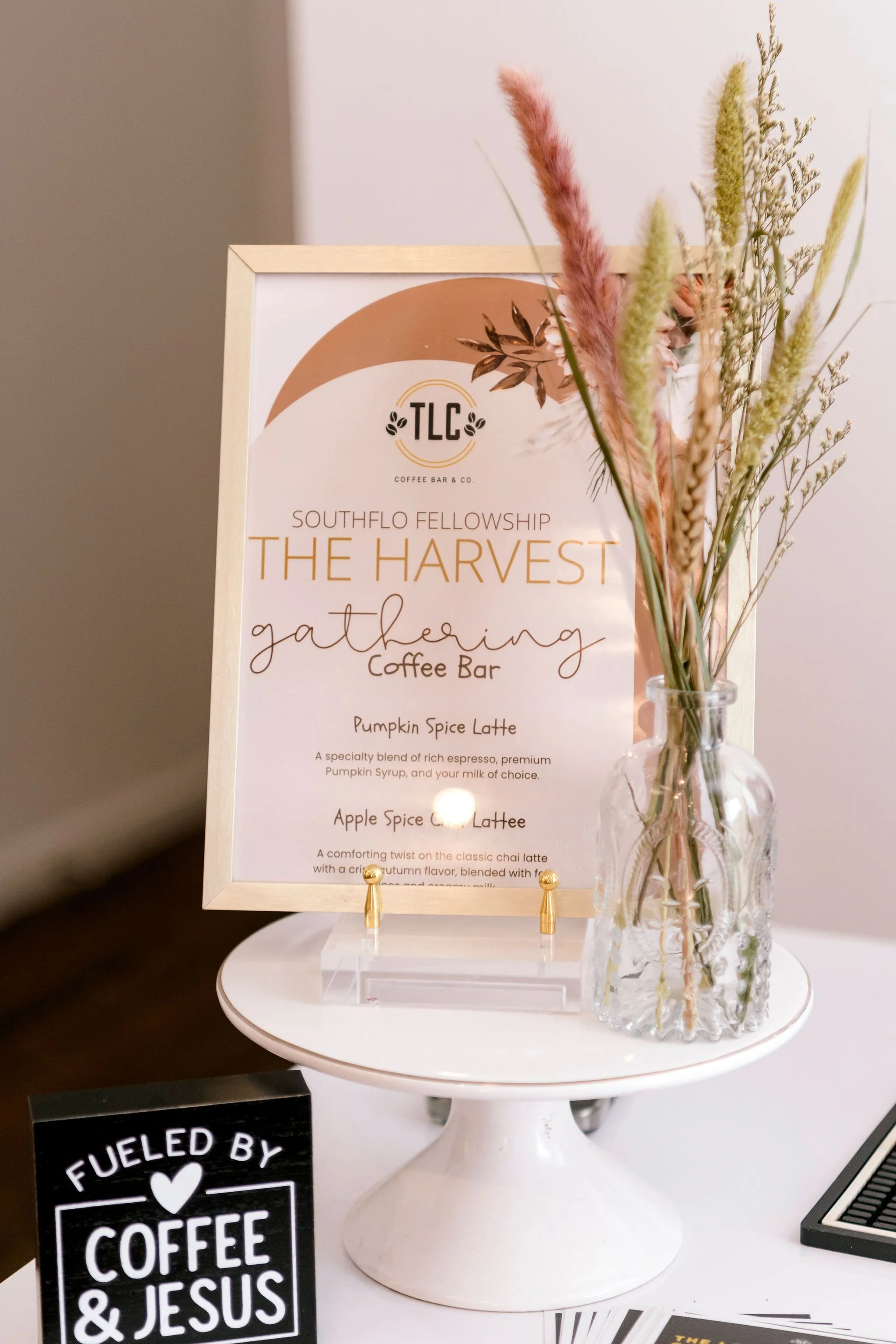 A framed sign promoting SouthFLO Fellowship's Harvest Gathering Coffee Bar, featuring Pumpkin Spice Latte and Apple Spice Chai Latte, with a small flower arrangement in a glass vase on a white table.