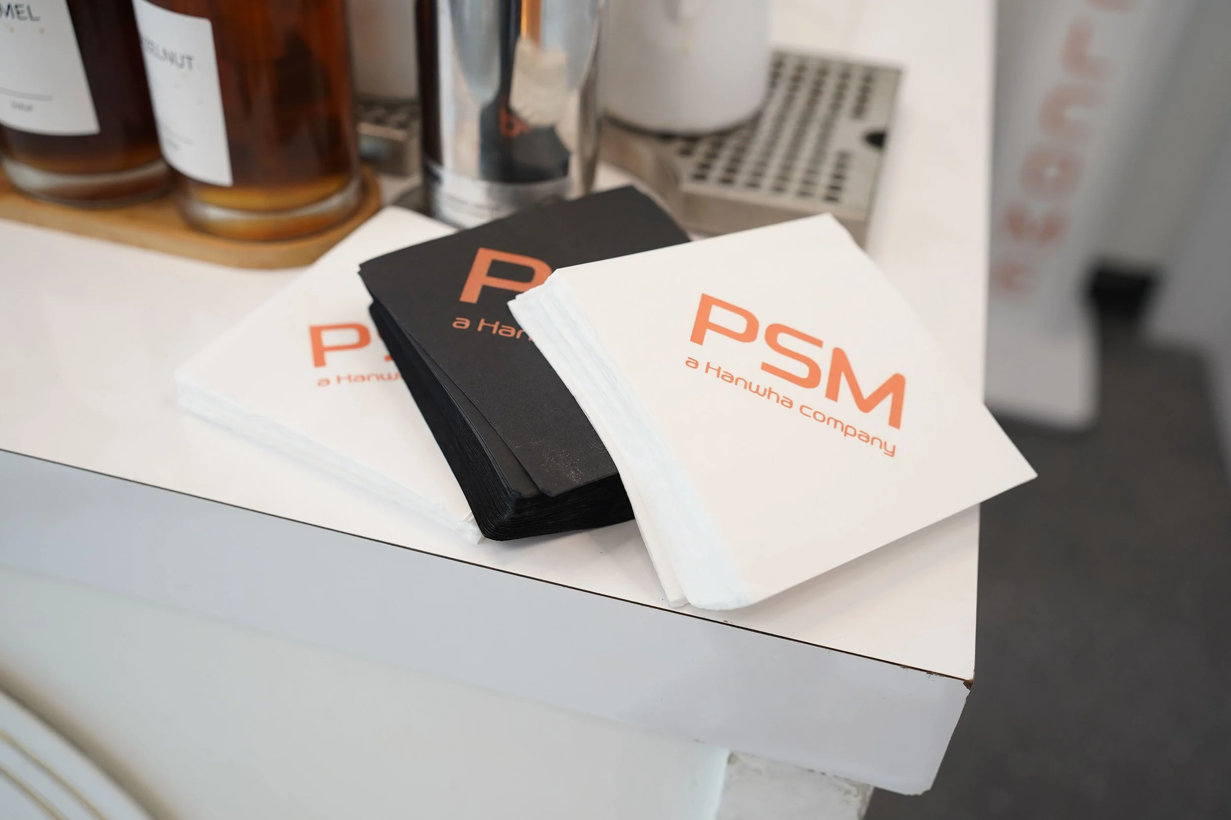 Stack of promotional papers with 'PSM a Hanwha company' printed on them, black and white, on a white counter with beverage bottles in the background.