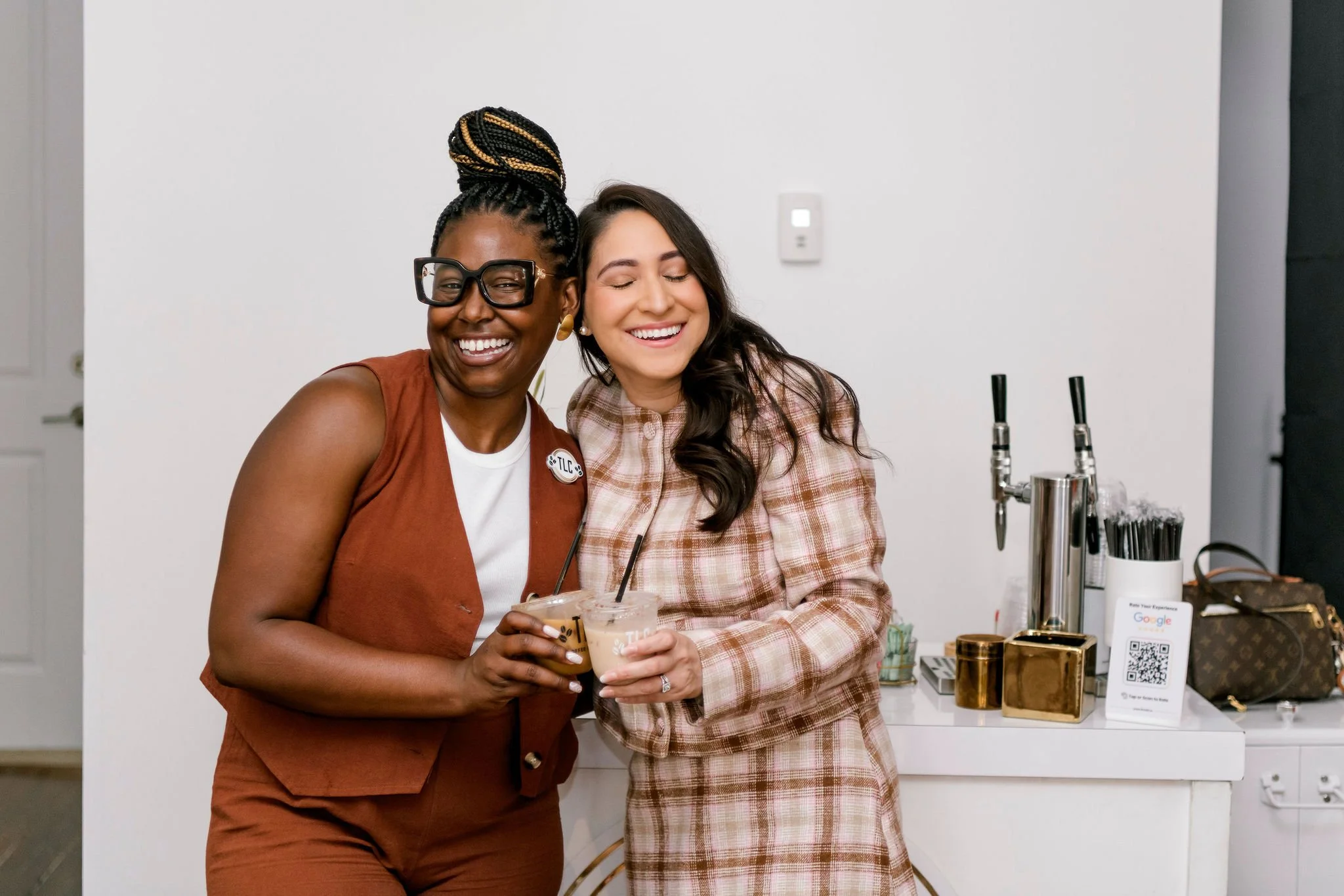 Two women smiling and holding iced coffee drinks at a social gathering or event, standing in front of a bar or counter with drinks, straws, and a QR code sign.
