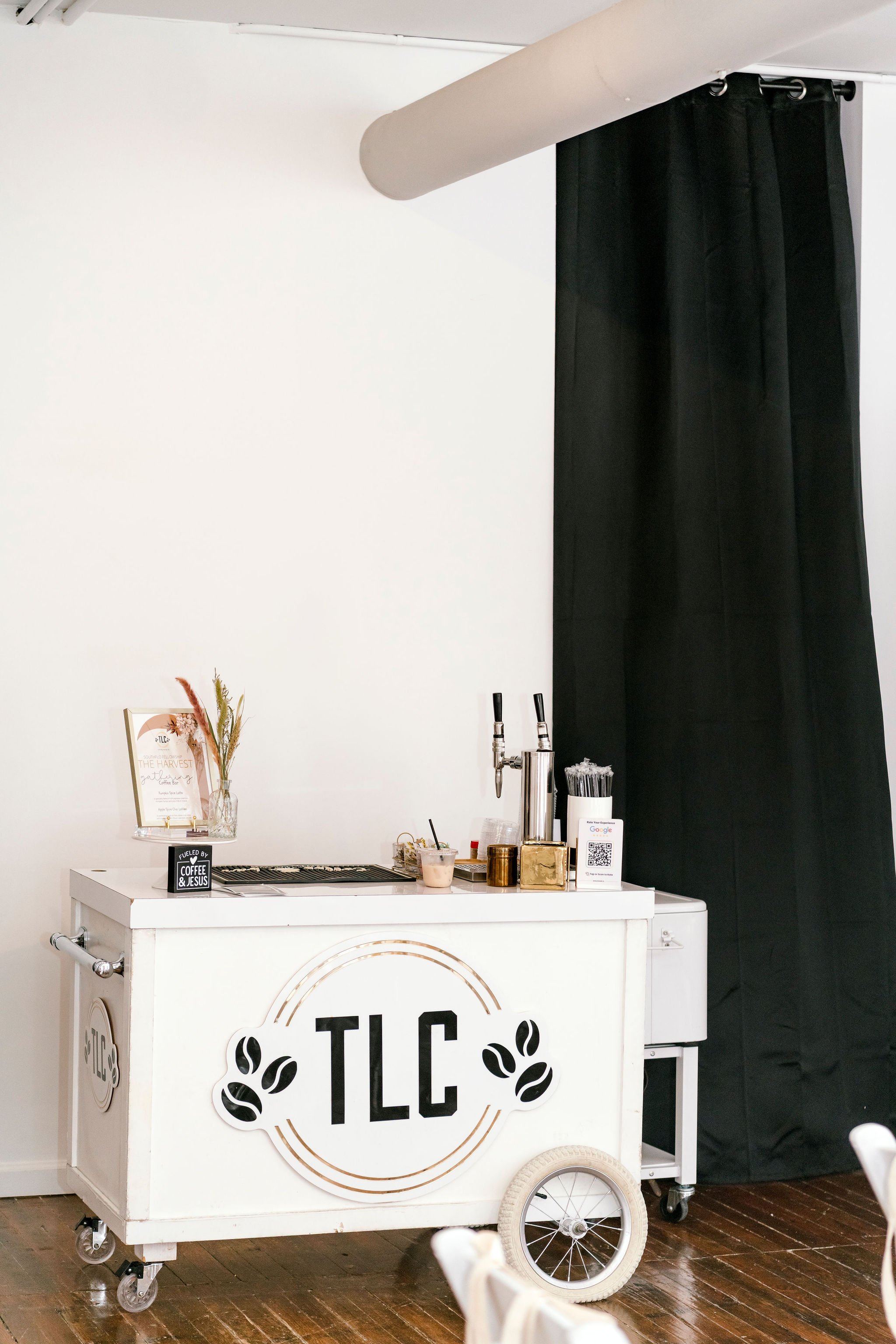 White coffee or beverage cart with TLC logo, black curtains, and barista equipment, including taps, on a wooden floor.