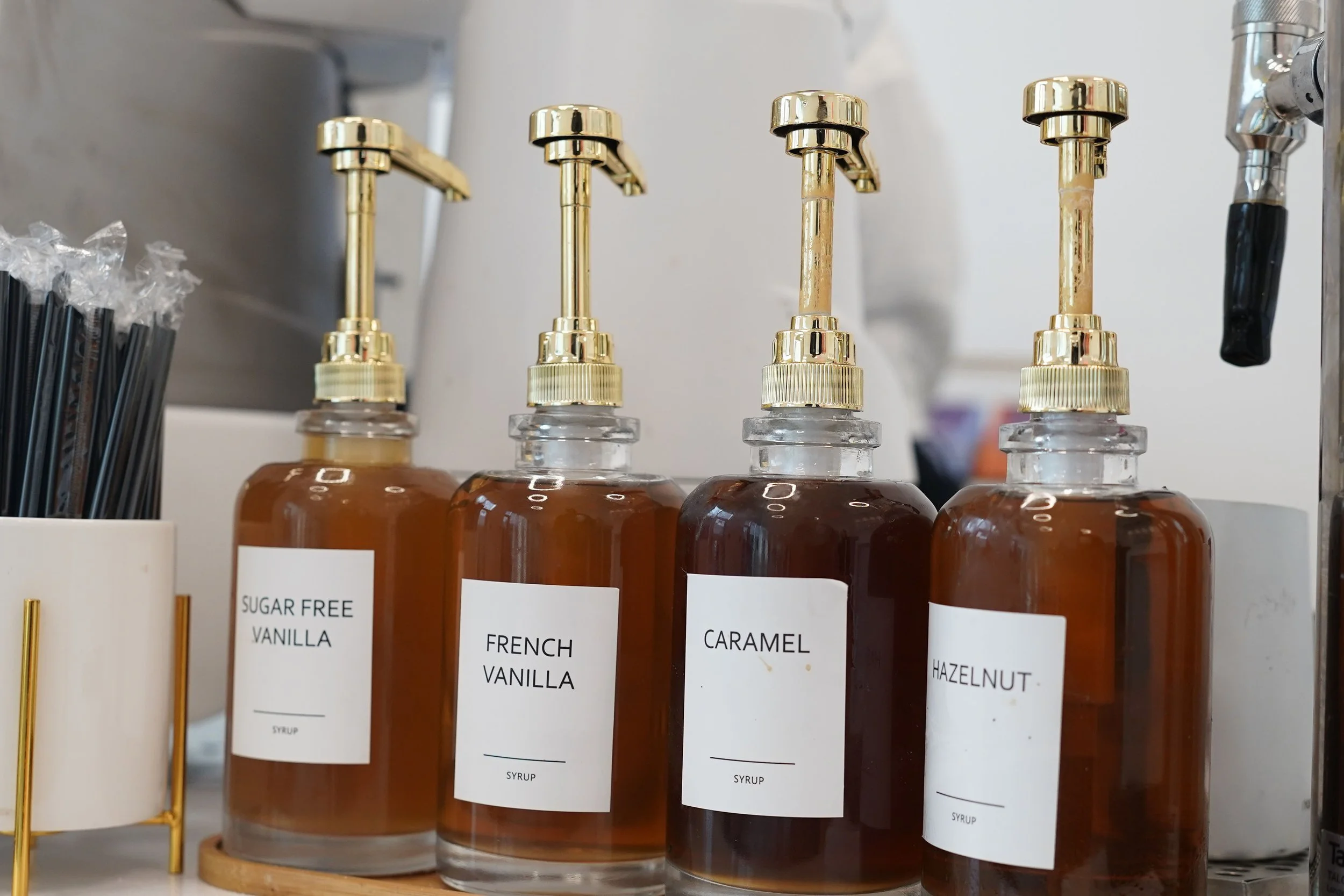Four glass syrup bottles with gold dispensers labeled 'Sugar Free Vanilla,' 'French Vanilla,' 'Caramel,' and 'Hazelnut,' placed on a round wooden tray.