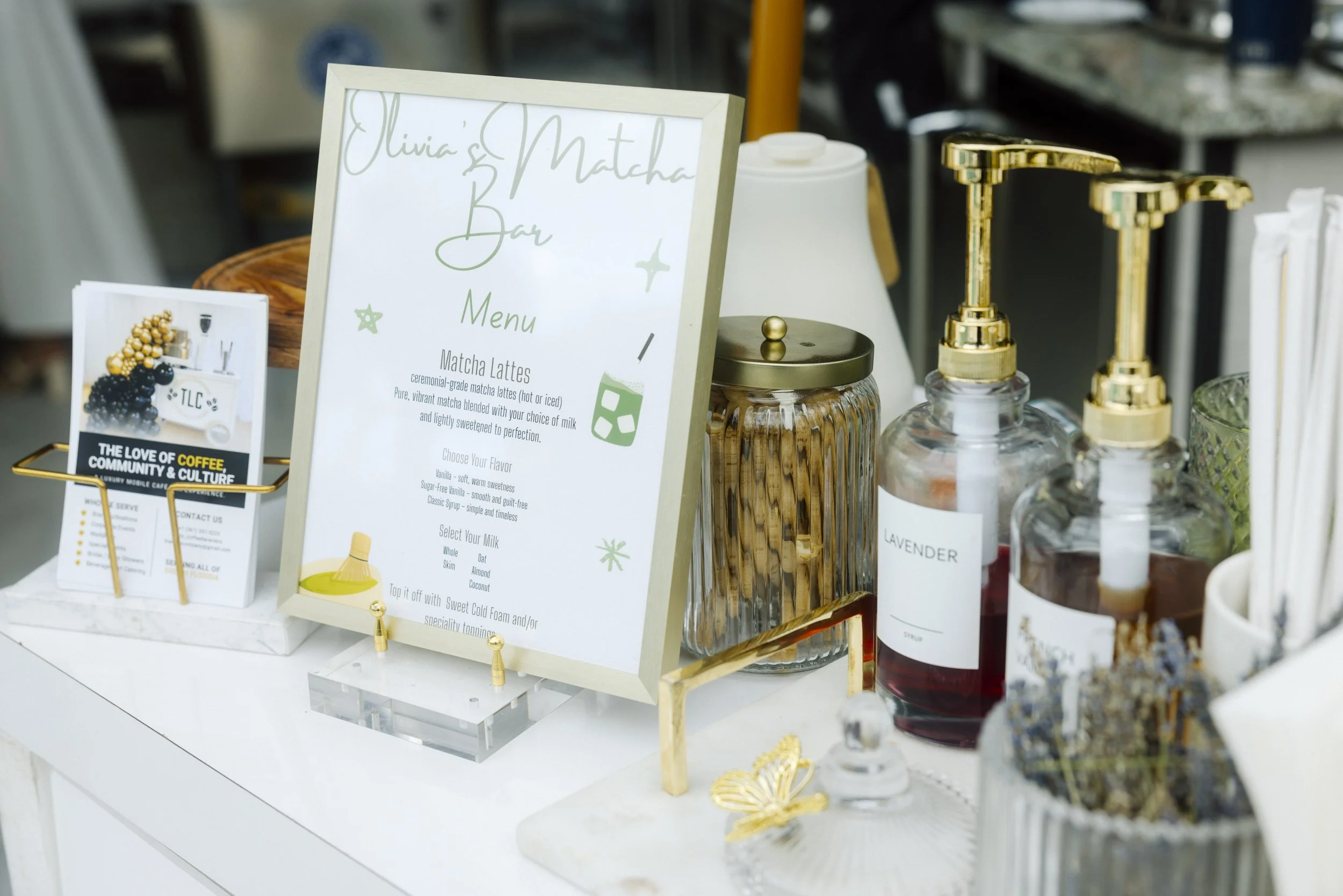 A menu sign for a matcha latte bar, with bottles of lavender syrup and other flavoring syrups, and decorative jars and items on a white counter. TLC Coffee Bar & Co. Premium Mobile Café Bar Experience for every event serving all of South Florida. Spe