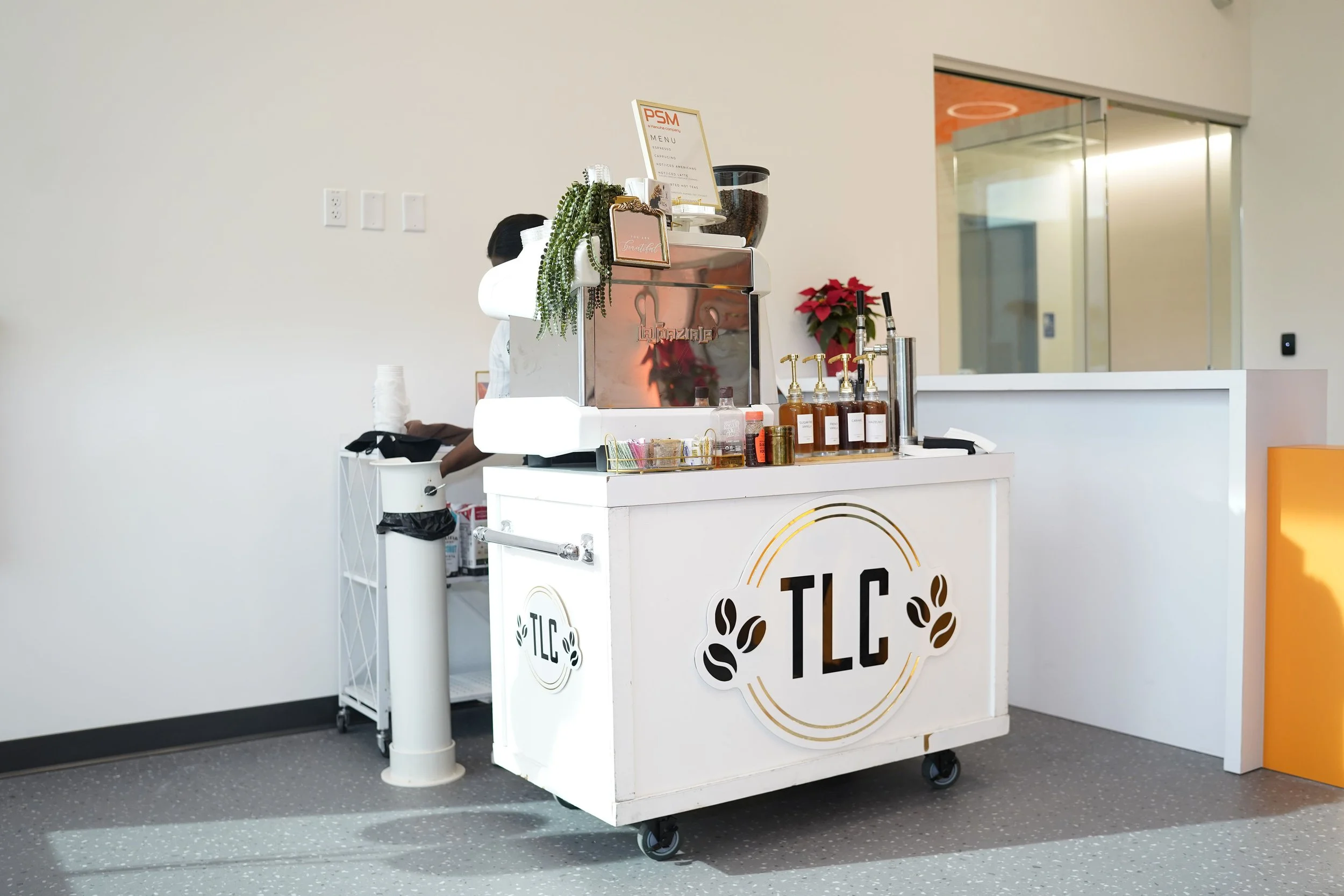 A small mobile coffee cart with the logo TLC and coffee bean designs, set up inside a building with a minimal background, featuring bottles of syrup, a coffee machine, and a poinsettia plant behind it.