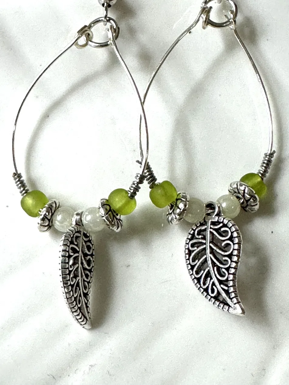 Handmade loop earrings featuring silver hoops, green and clear glass beads, and a detailed silver leaf charm — part of the Moss Light collection. Hypoallergenic ear wires.