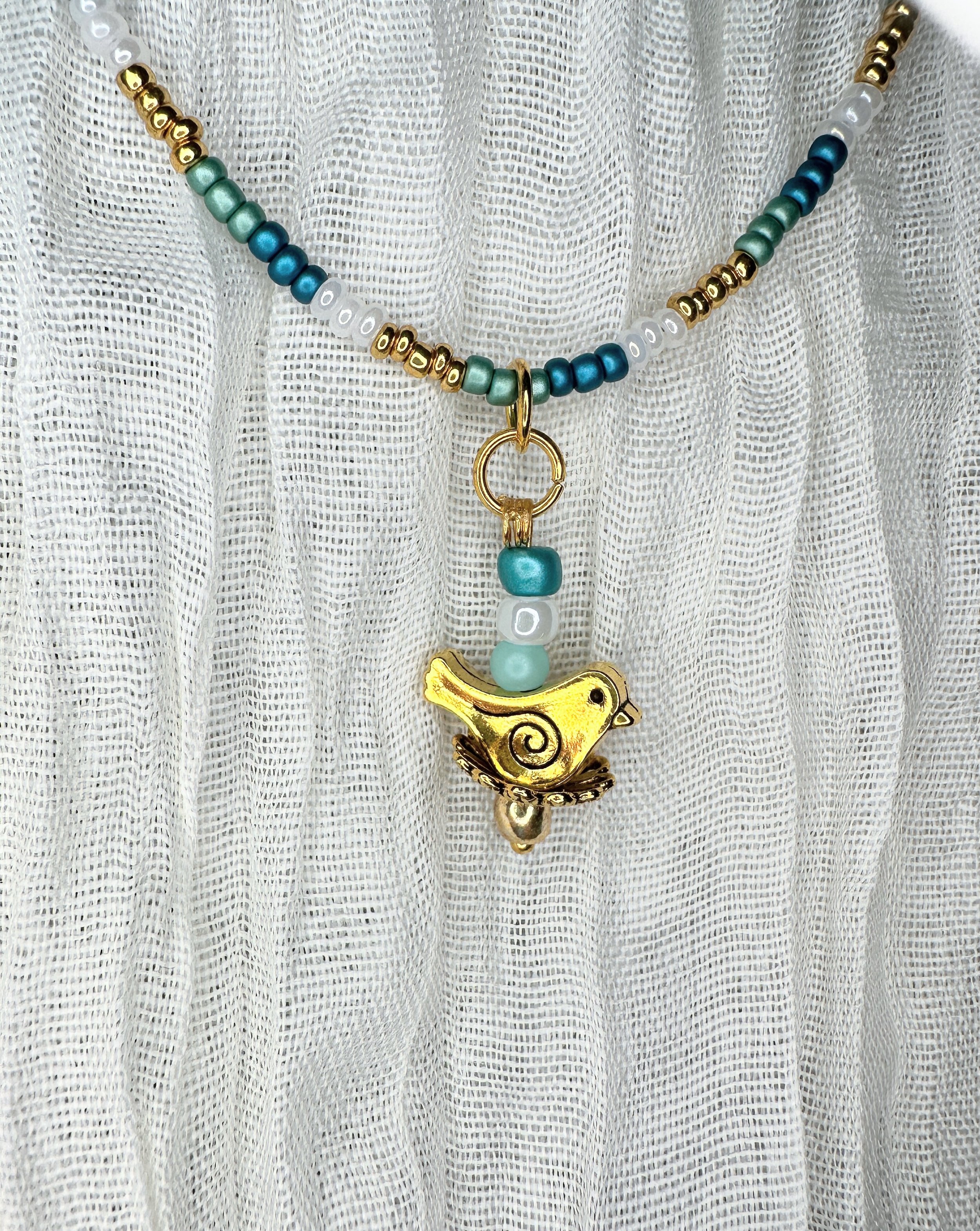 Handmade children's necklace featuring a gold bird charm with teal, gold, and white glass beads — part of the Bluebell collection. Hypoallergenic hardware, hand-strung.