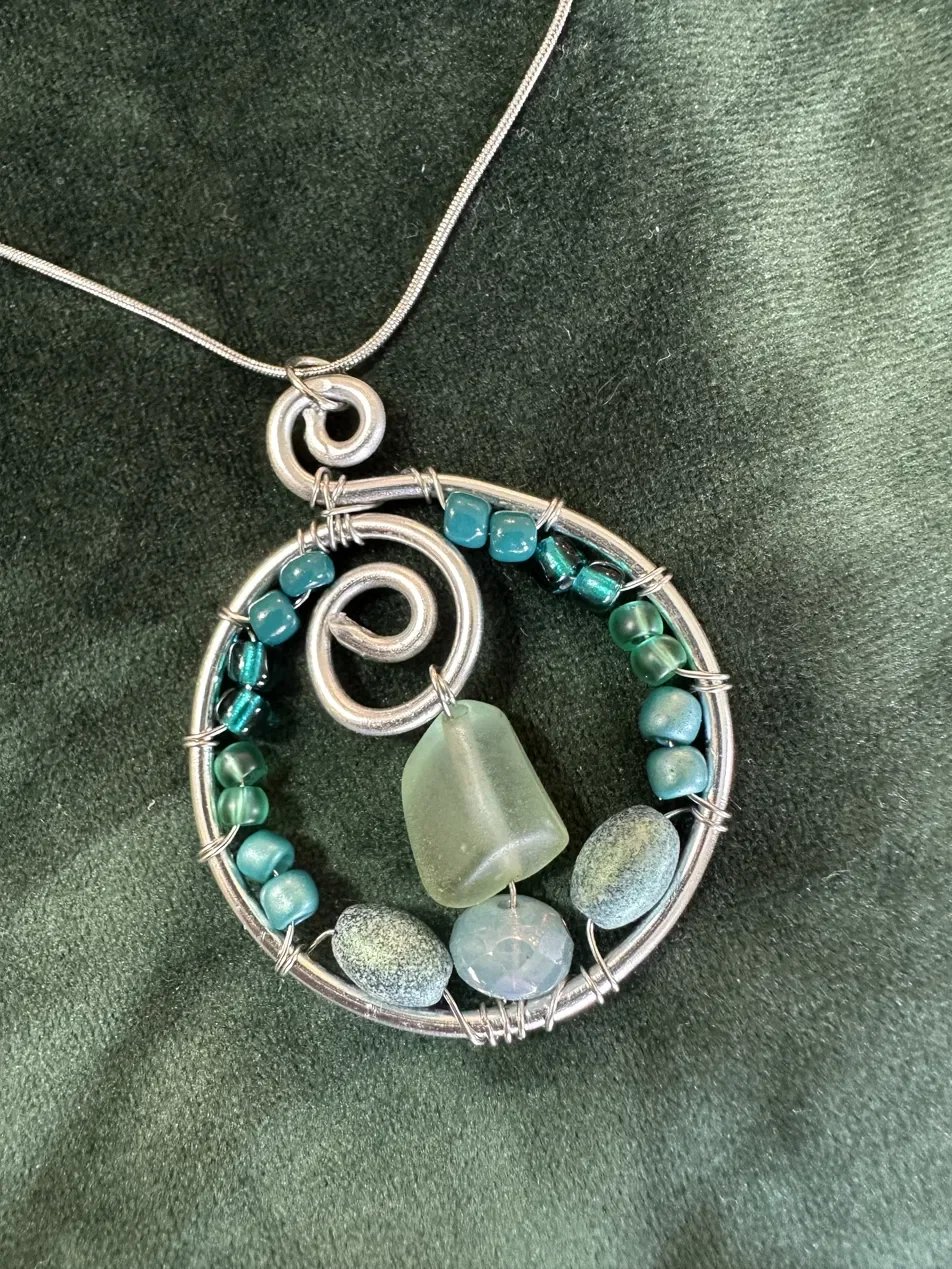 Handmade signature necklace featuring a hand-formed silver spiral pendant, sea glass, and teal glass beads — part of the Still Water collection. Wire-sculpted and finished with hypoallergenic hardware.