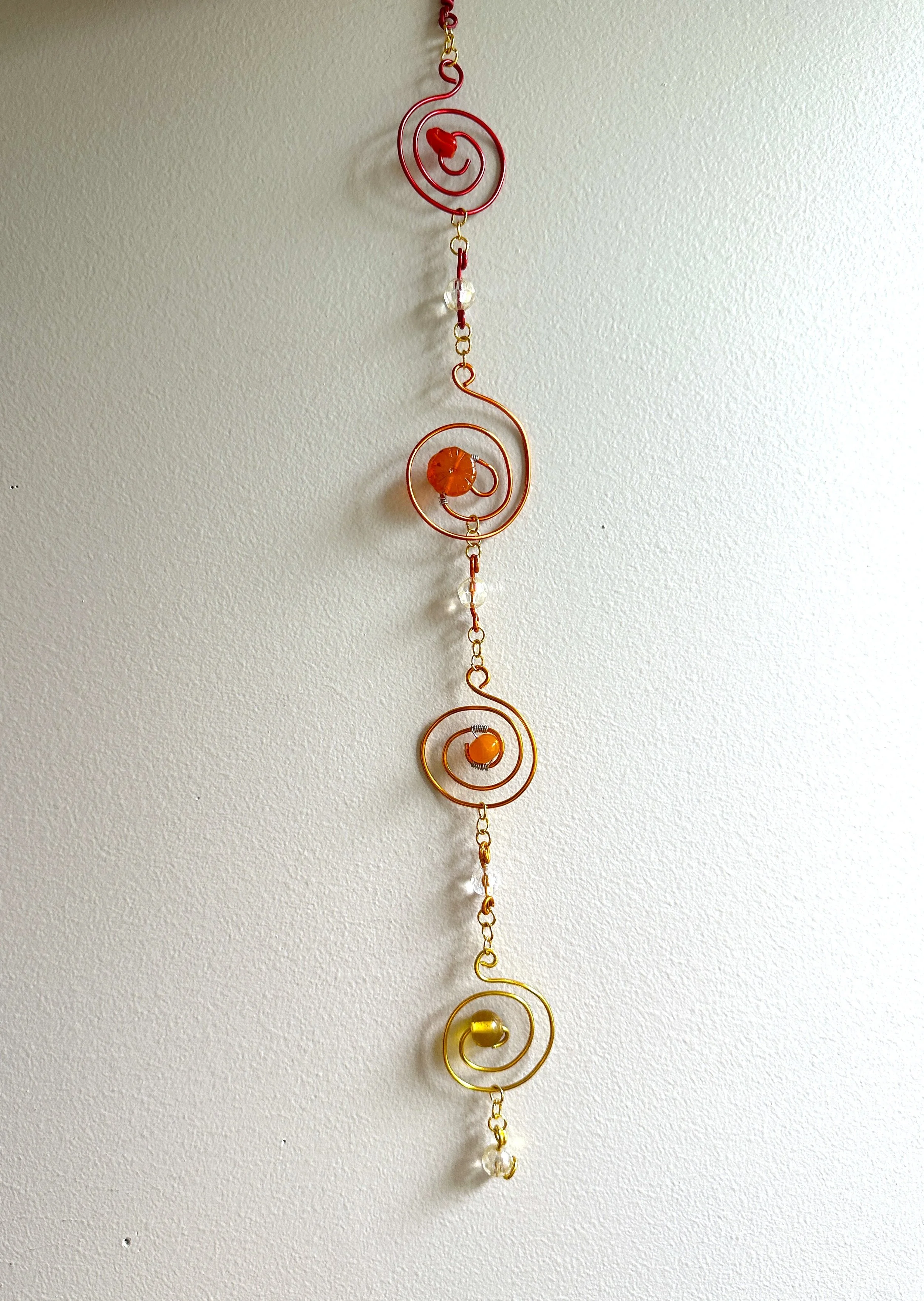 A wire-wrapped spiral sun catcher in sunny red, orange, and gold hues with glass bead accents.
Part of the Hello Summer line — bright, warm-toned pieces meant to catch sunlight and celebrate the season.