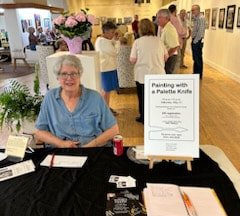 Woman sitting at a table with a sign about a painting event, surrounded by people in an art gallery.