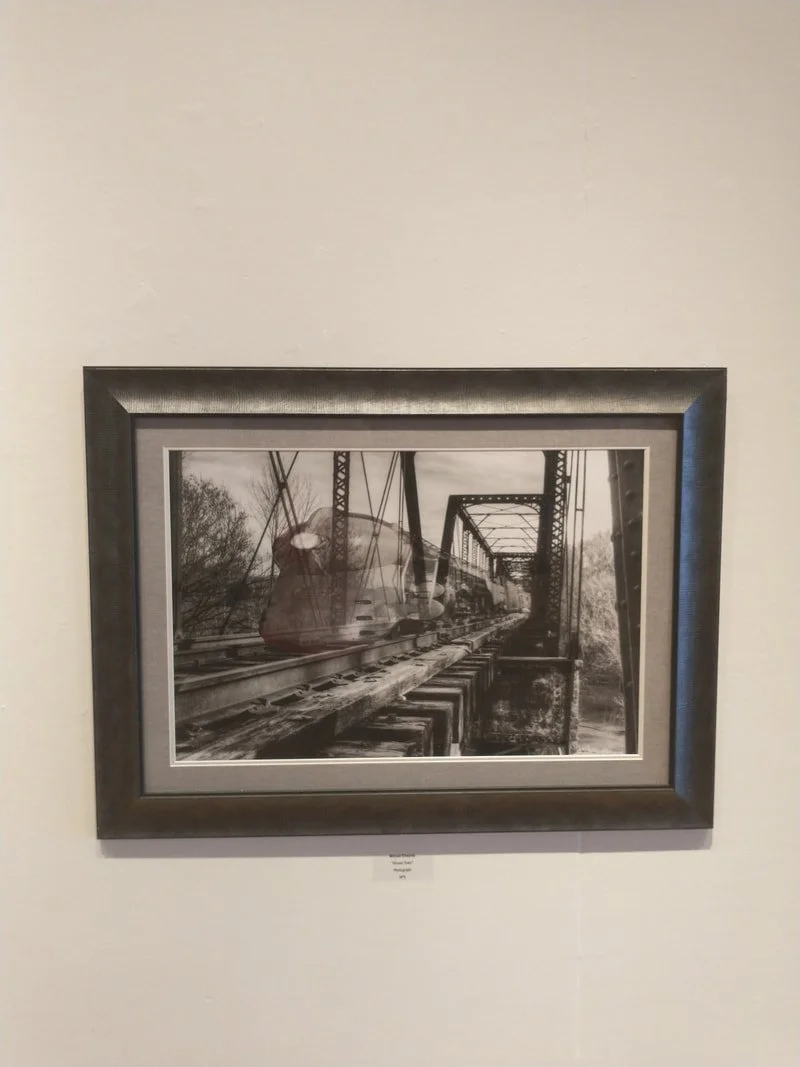 Black and white photograph of a train on a bridge with trees in the background, framed and hung on a white wall.