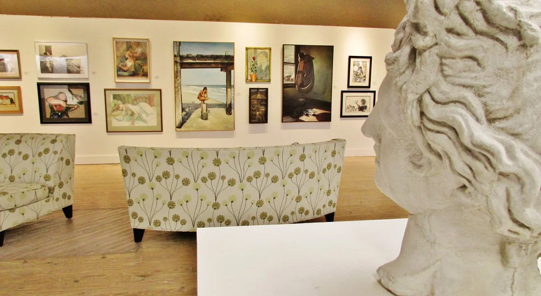 Museum gallery with multiple paintings on the wall, two floral-patterned sofas, and a bust sculpture in the foreground.