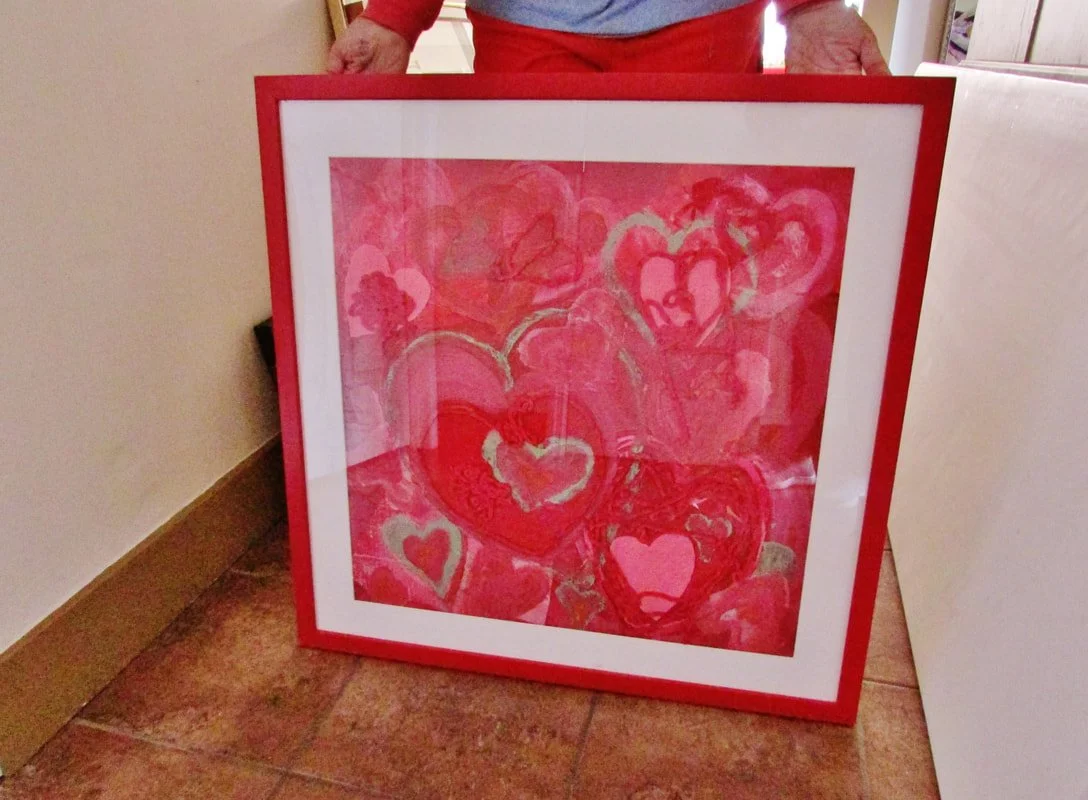 A framed painting of multiple pink and red hearts, held by a person wearing a red shirt and blue apron, standing on a tiled floor.