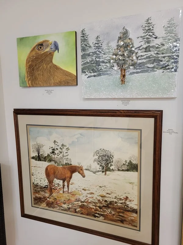 Three paintings on a wall: a close-up of a hawk's head, a snowy landscape with a decorated tree, and a horse standing in a field with trees in the background.