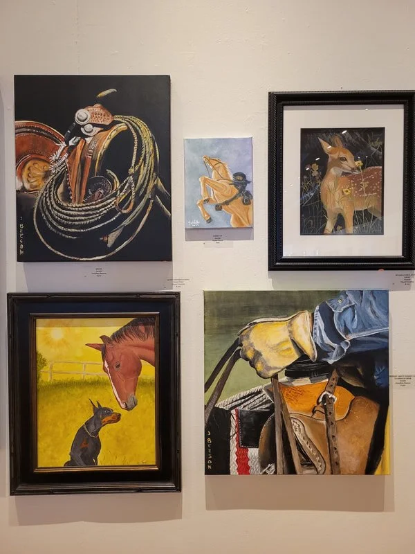 A gallery wall with five paintings including a lasso and horse, a woman in a yellow dress, a deer, a horse with a dog, and a close-up of a person’s hand holding a saddle.