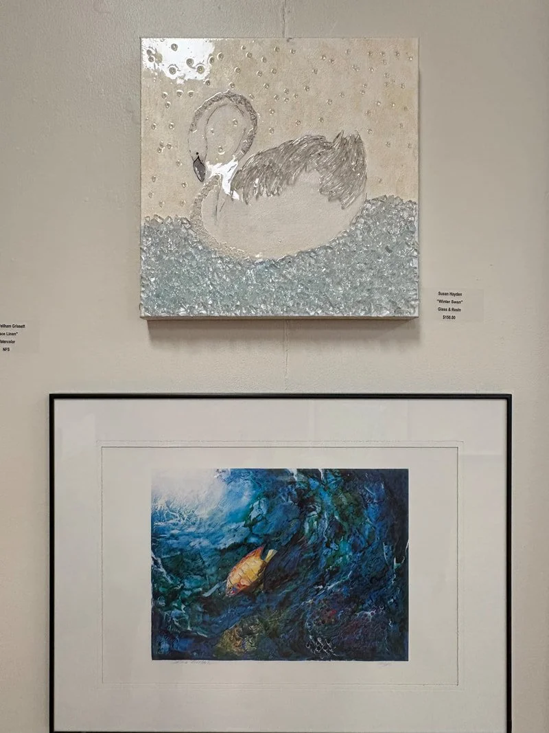 An art display with two paintings. The top painting depicts a swan with a blurred face and a textured background in shades of white and silver. The bottom painting shows an abstract underwater scene with a fish and swirling blue and green colors.