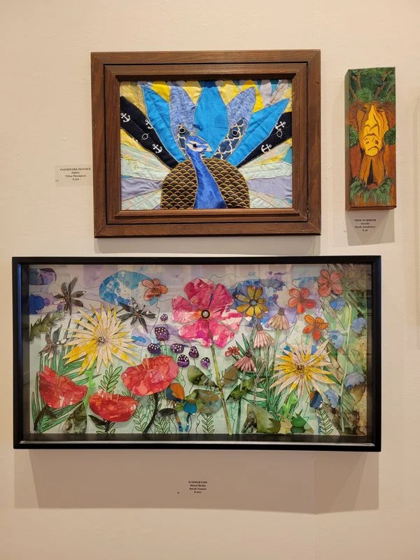Three colorful artworks displayed on a wall: a framed fabric collage of a peacock with spread tail feathers, a rectangular piece depicting a tiger's face surrounded by greenery, and a watercolor-style painting of various flowers and insects in a gard