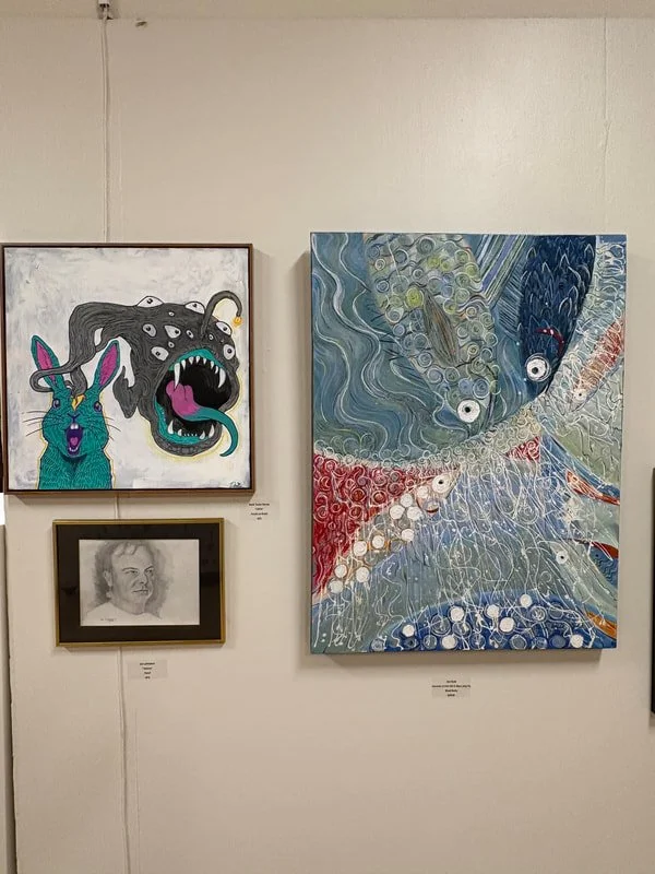 Gallery wall with three artworks: a colorful painting of a creature with a long tongue, a surreal abstract painting with swirling patterns and multiple eyes, and a black-and-white portrait drawing of a person.
