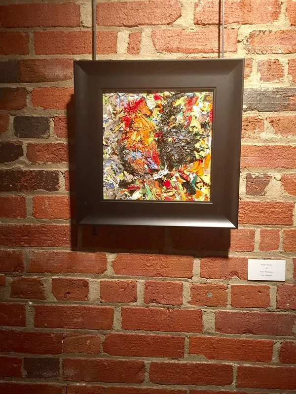 Abstract textured painting with vivid orange, red, yellow, black, and white colors, framed in black and hung on a brick wall.
