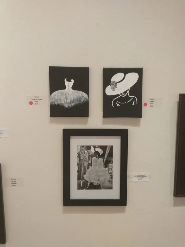 Three pieces of artwork on a gallery wall: two black and white paintings at the top and a black-and-white photograph in a frame below. The left painting shows two figures standing on a fuzzy, textured surface. The right painting depicts a woman weari