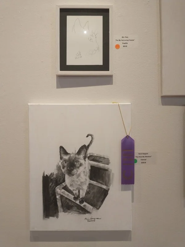 Black and white charcoal drawing of a cat sitting in a small box with a wooden plank and a branch-like object behind it, displayed in an art gallery.