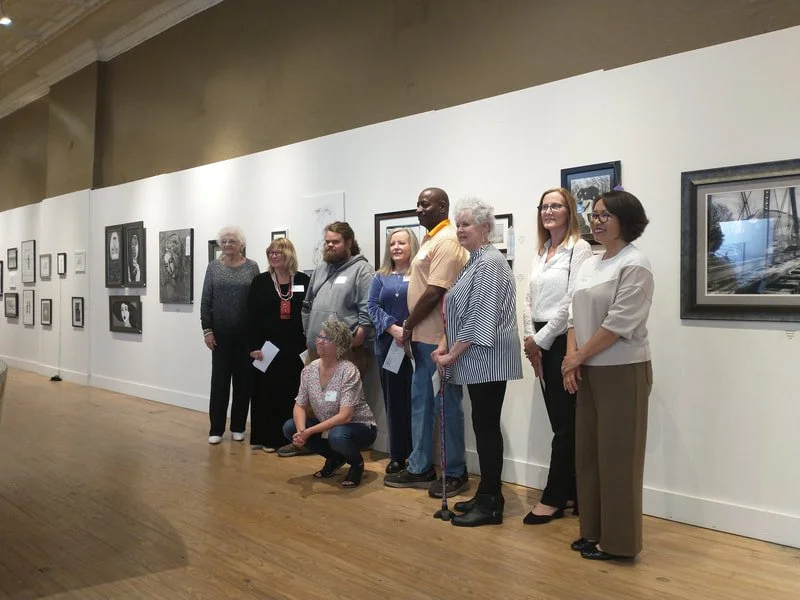People standing and sitting in an art gallery with framed artworks on the wall.