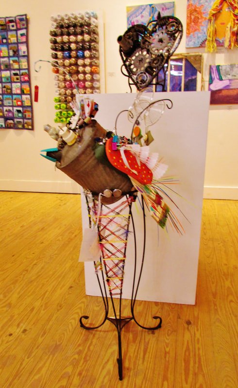 A whimsical sculpture resembling a person created from various crafting and art supplies, standing on a wooden floor with colorful artwork displayed on the walls behind it.